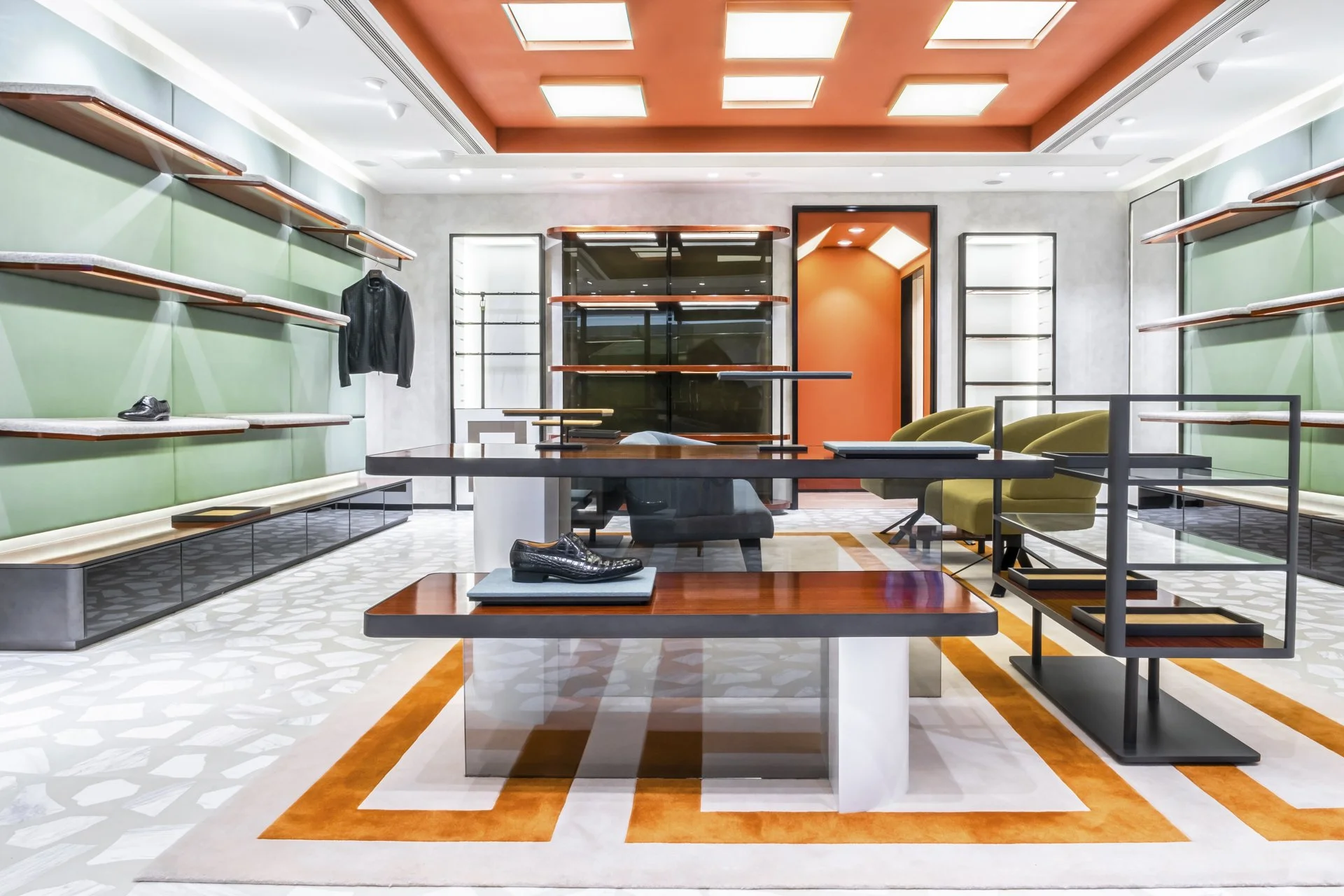 A modern retail store interior with light green walls, black shelving, and a large central table displaying men's shoes. There are yellow chairs, glass display cases, and a decorative orange and white rug on a geometric patterned floor. The ceiling f