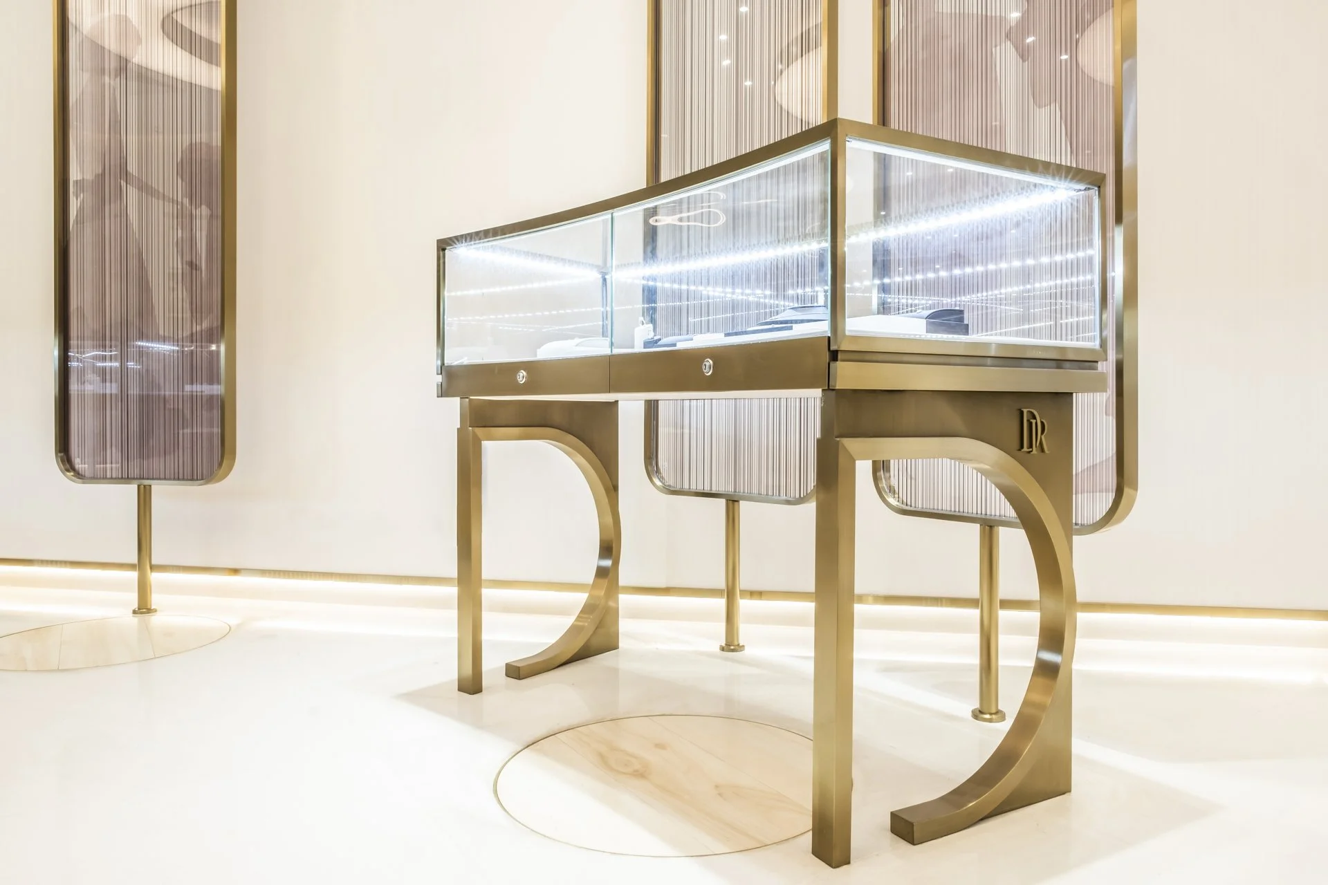 A modern, luxurious display case with glass sides and gold metal frame, featuring LED lighting inside. The case is positioned in a gallery-like setting with a cream-colored wall behind it and decorative panels with a gold tint on either side.