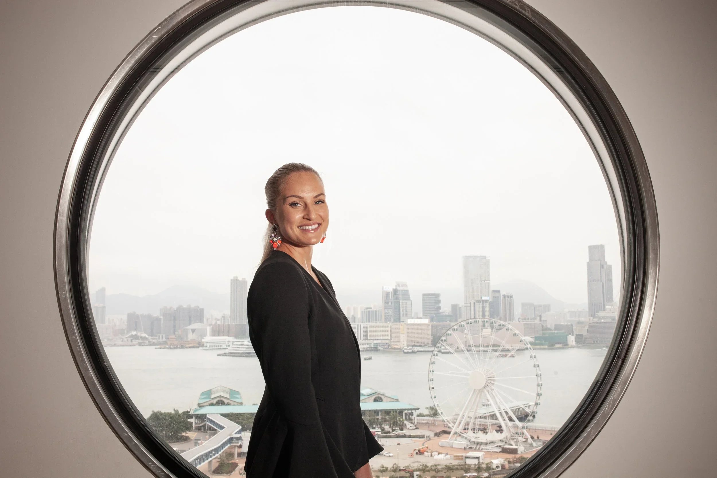 A woman with blonde hair tied back, wearing earrings and a black blazer, stands in front of a large circle window overlooking a city skyline with various high-rise buildings, a Ferris wheel, and a body of water.