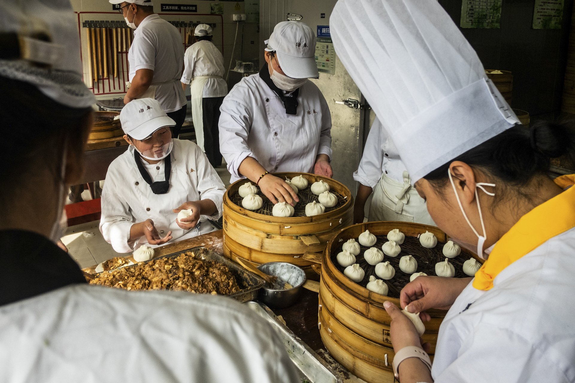 Several people wearing white uniforms, masks, and hats are preparing and filling steamed buns in a kitchen or restaurant setting. Steamed buns are placed in bamboo steamers on the table.