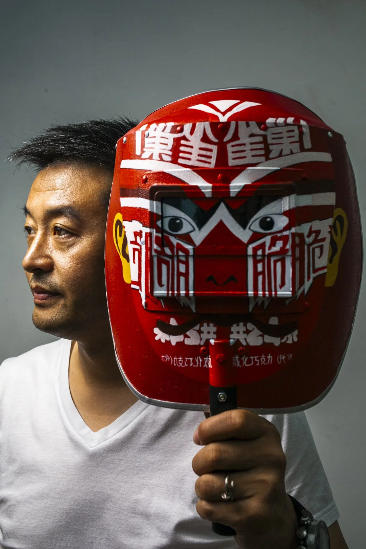 A man with short black hair and wearing a white t-shirt is holding a red and black Chinese-style decorative face mask on a stick in front of his face. The mask has a stylized lion face with bold colors and traditional Chinese characters and design el