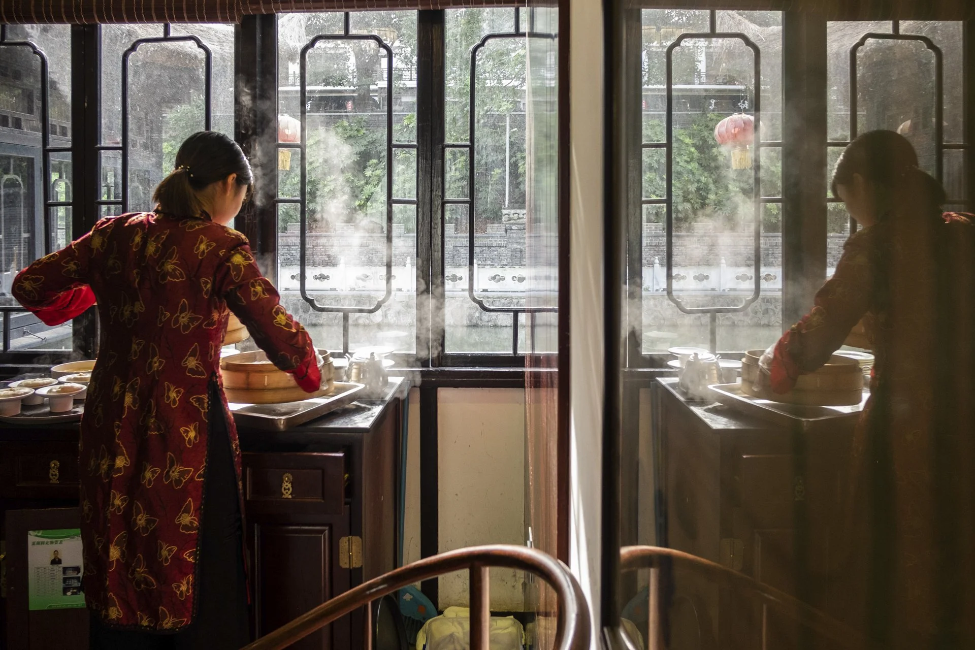 A woman in a red dress with butterfly patterns is standing in front of a window in a restaurant or kitchen, preparing food at a steam table with multiple bowls. There is steam rising from the food, and outside the window, leafy trees and some red lan