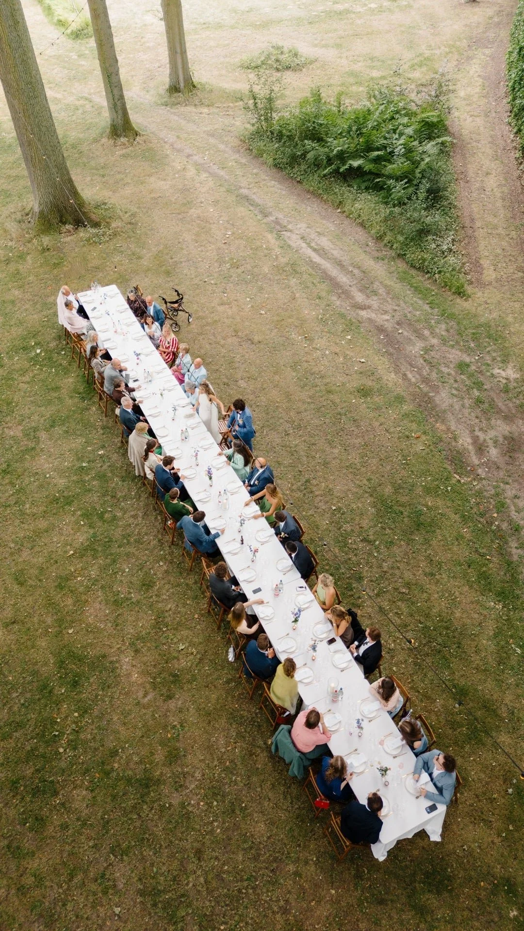 Langgerekt restauranttafels met mensen die aan een buitengelegen evenement deelnemen, met witte tafelkleden en bloemen, in een groen park.