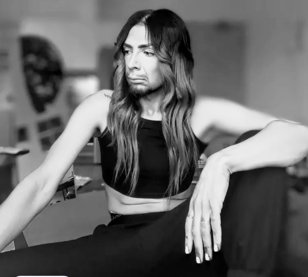 Black and white photo of a person with long wavy hair, wearing a sleeveless crop top, sitting with one arm resting on a surface and looking contemplative.