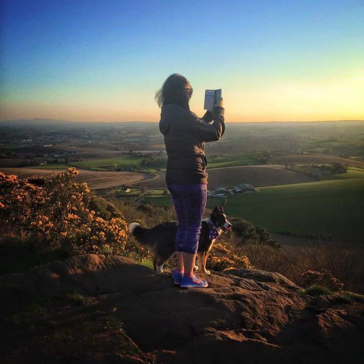 Landscape photo of a sunset in Scrabo Country Park in Northern Ireland with Joanna Ritchie taking a photograph on her phone.