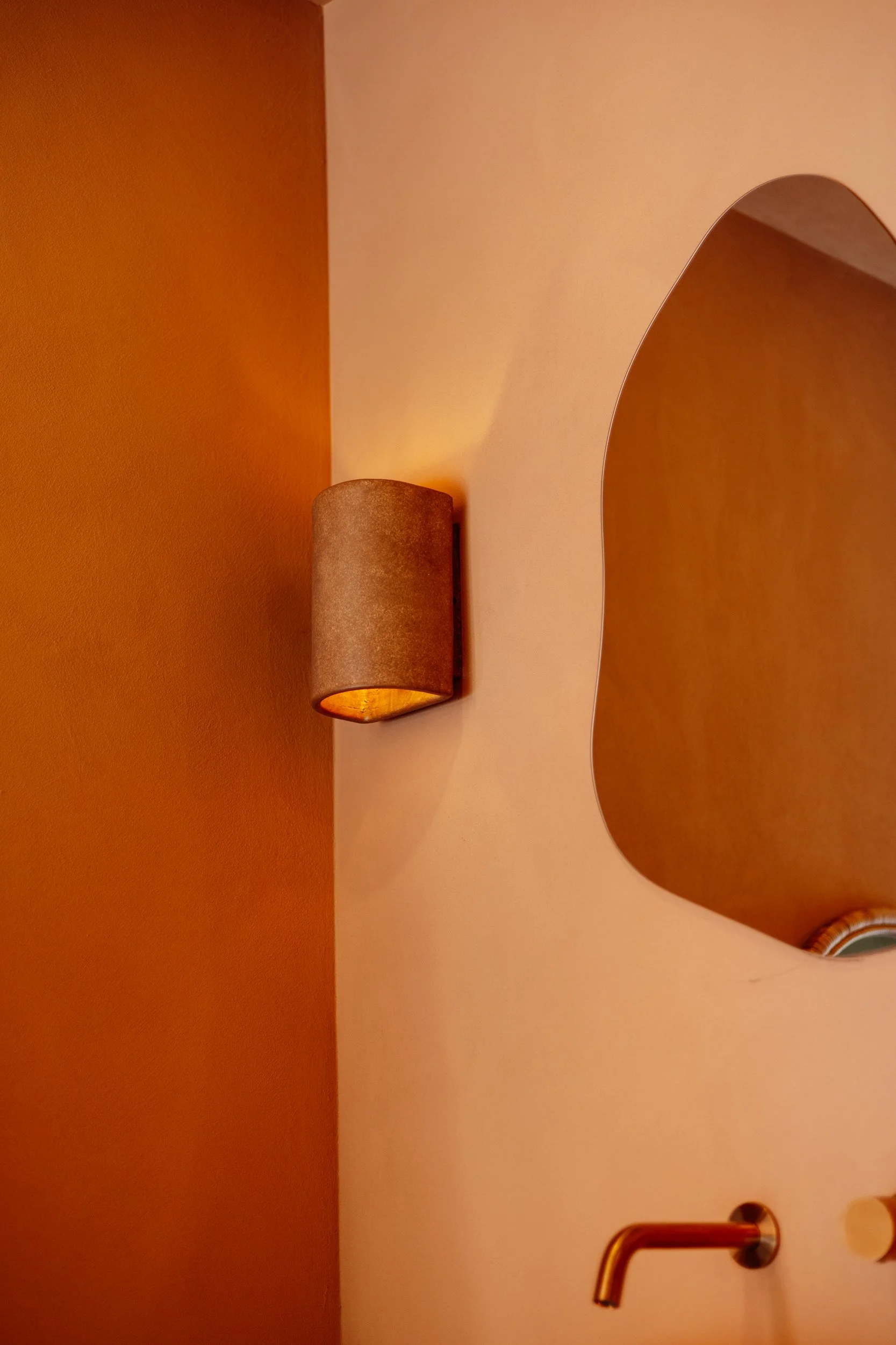 Terracotta bathroom wall with an irregular mirror and cylindrical sconce at Stepney Warehouse