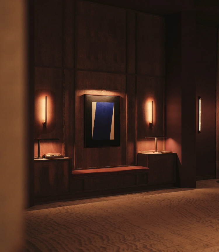 A warmly lit wooden-paneled wall with a central framed abstract artwork, two wall-mounted lamps on either side, and two side tables with lamps and books.