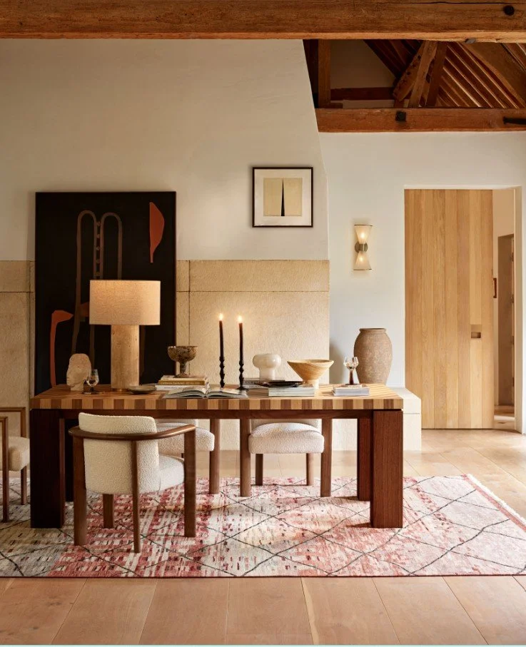 A dining room with a wooden table set with decorative items, candles, and books. There are beige upholstered chairs, a patterned rug, and abstract artwork on the wall.