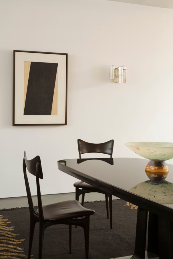 Luxury dining room with a black table, two black chairs, a wall-mounted glass light fixture, a framed abstract geometric art piece from artists Studio Earle