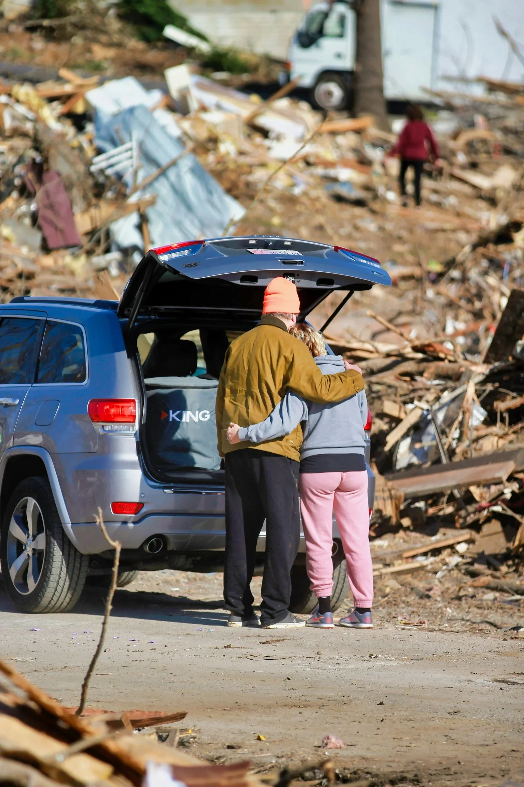 A couple stands embraced next to a blue SUV with its trunk open, amidst debris and rubble, likely from a natural disaster. Another person walks in the background, surveying the damage.