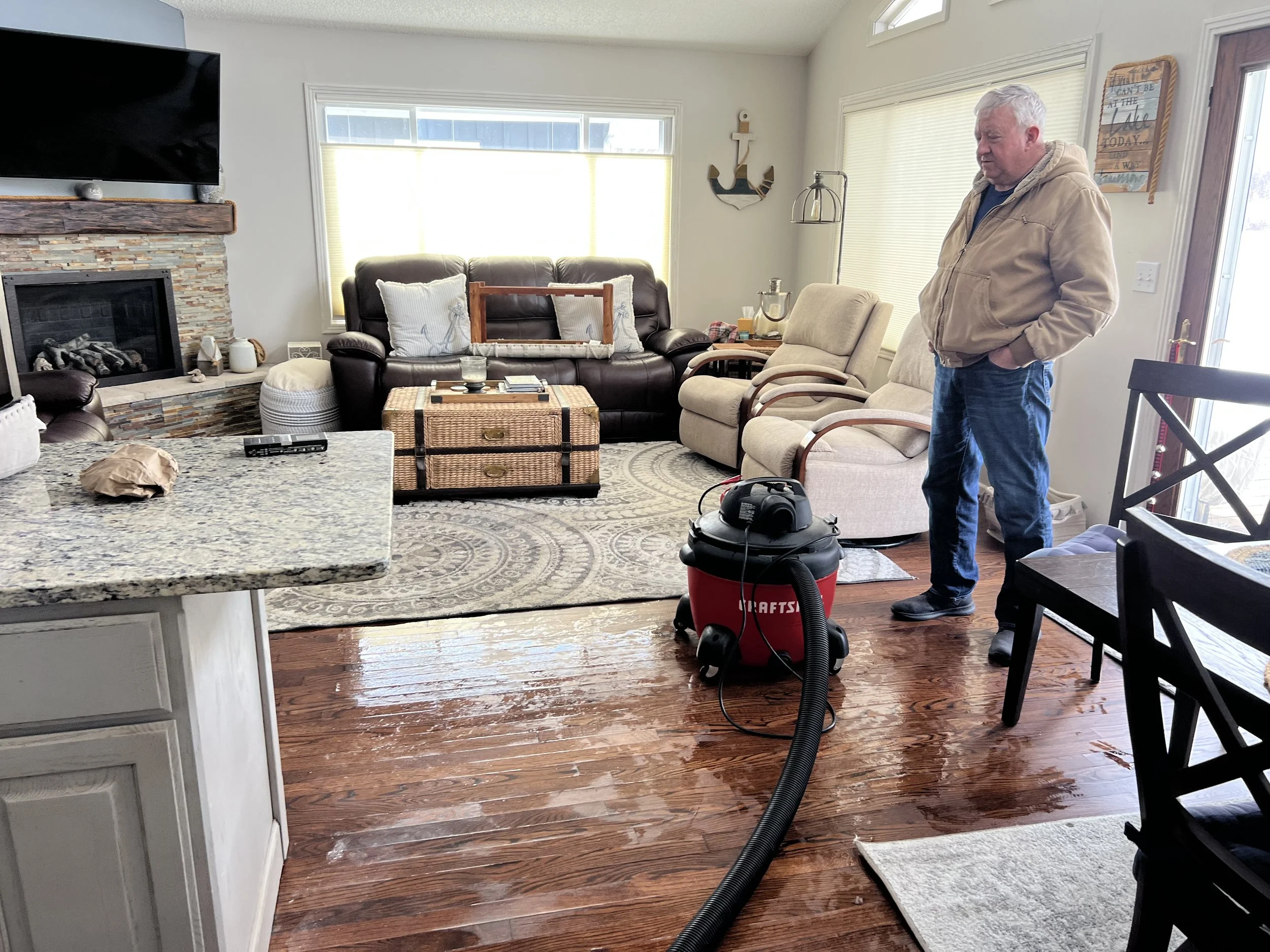 A person standing in a living room with hardwood floors, a vacuum, and some water on the floor. The room has a sofa, recliner, TV, and a fireplace.