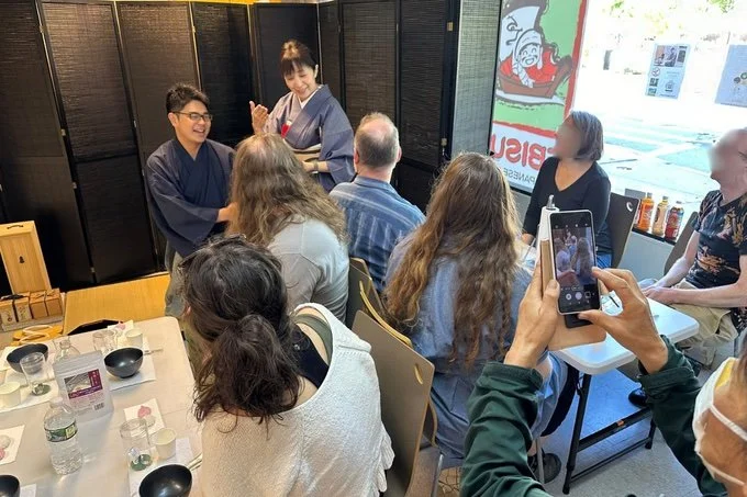 People gathered in a restaurant, sitting around tables, watching a Japanese chef preparing food, with some taking photos, and a window showing outside street view.