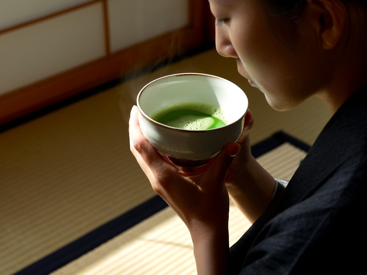 A person holding a white bowl with steaming green tea in a traditional Japanese room with tatami mats.