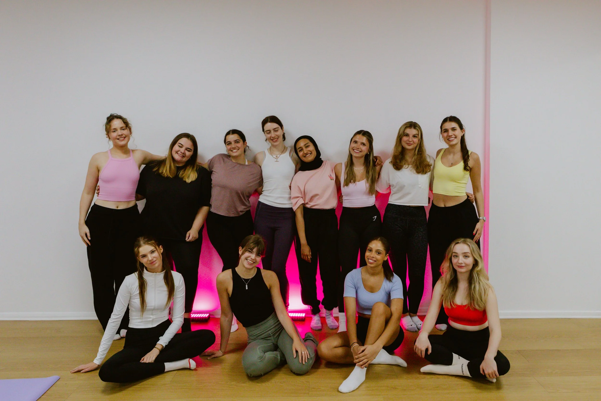 Group of young women in activewear posing in a fitness class studio with a white wall and pink lighting.