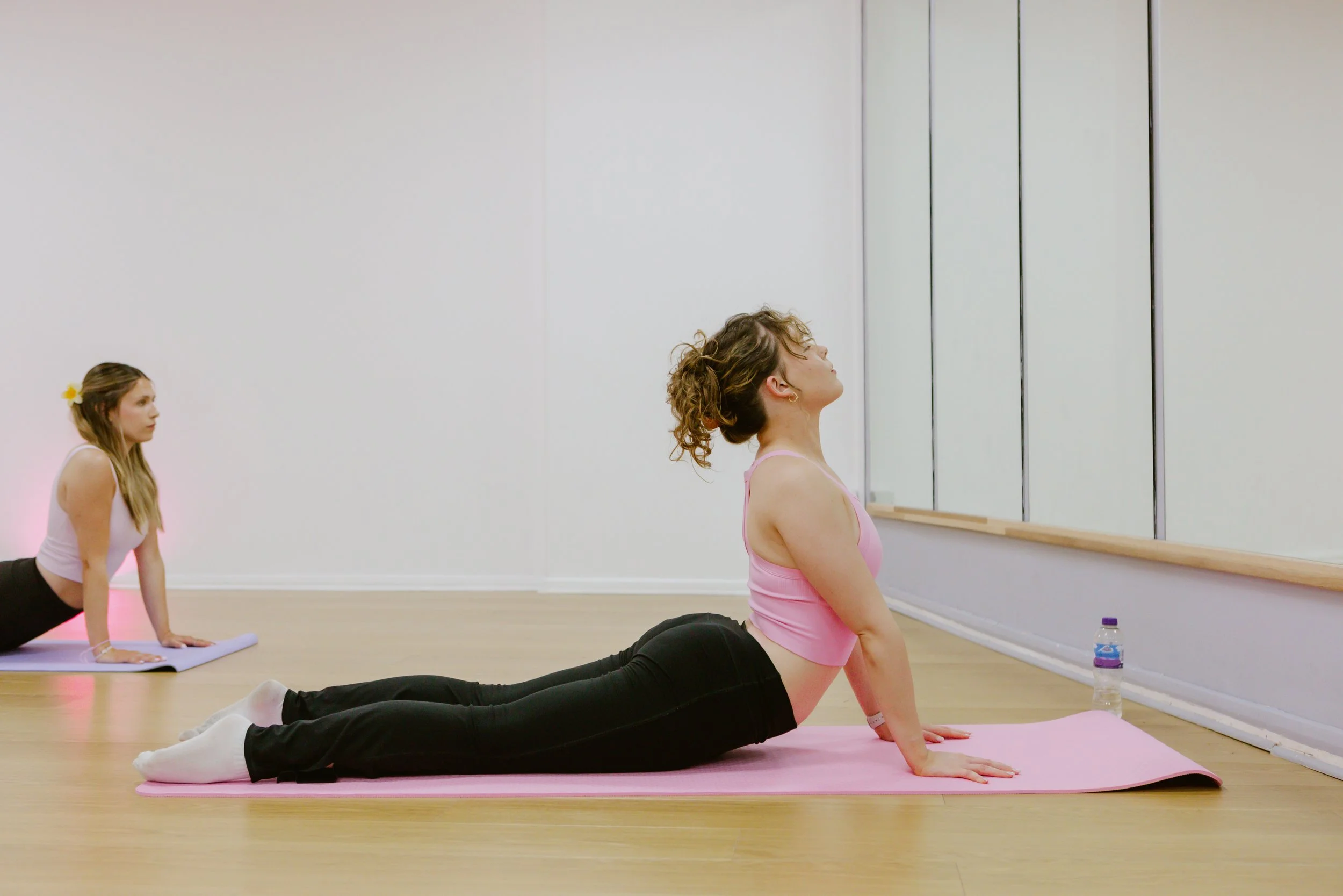 Two women practicing yoga in a studio, one woman is in a cobra pose, and the other woman is in a plank pose, facing a mirror.
