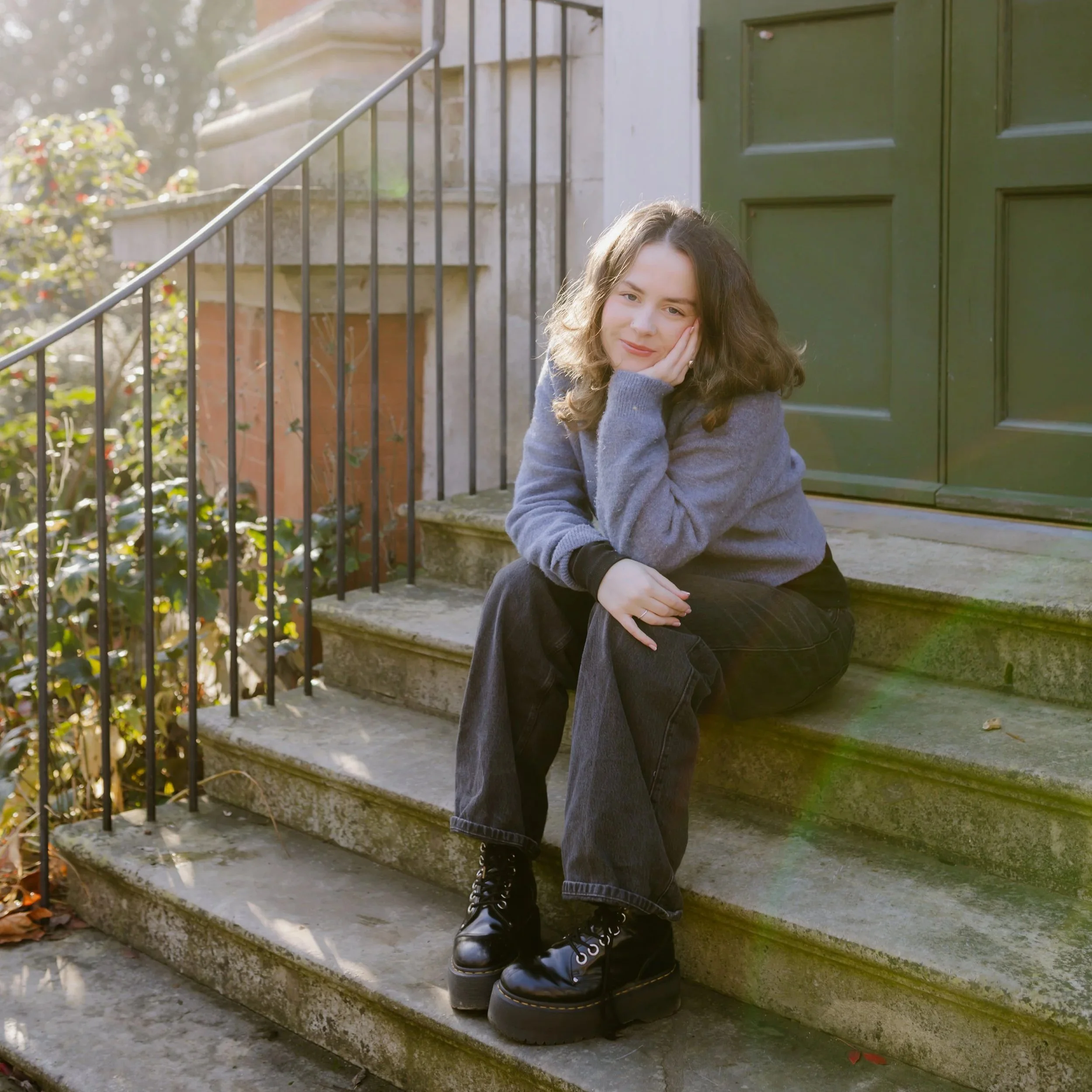 A woman with long, wavy brown hair and light skin is sitting on the steps outside, resting her face on her hand and smiling softly. She is wearing a gray sweater, black pants, and black combat boots. The steps are made of concrete, and there is a green door behind her. To her left, a black metal railing runs along the stairs, and some plants and leaves are visible in the background.