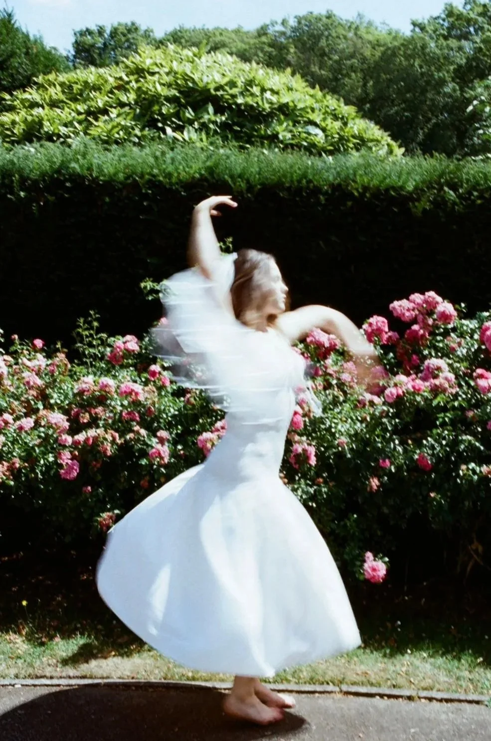 A woman in a white dress dancing barefoot among pink roses and green bushes in a garden.