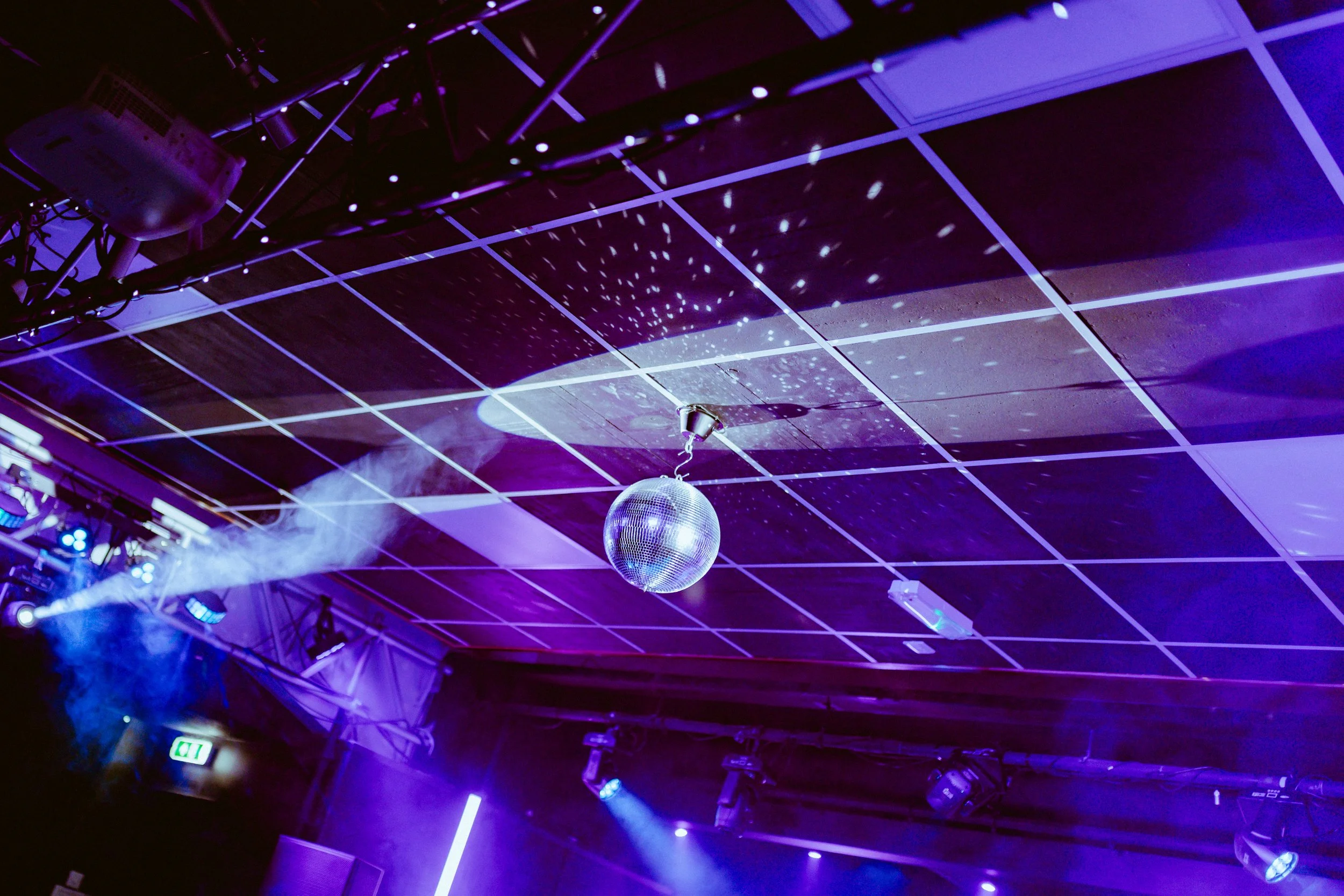 A ceiling with grid tiles illuminated by purple and blue stage lighting, a disco ball hanging from the ceiling, and some fog effects in a nightclub or event space.