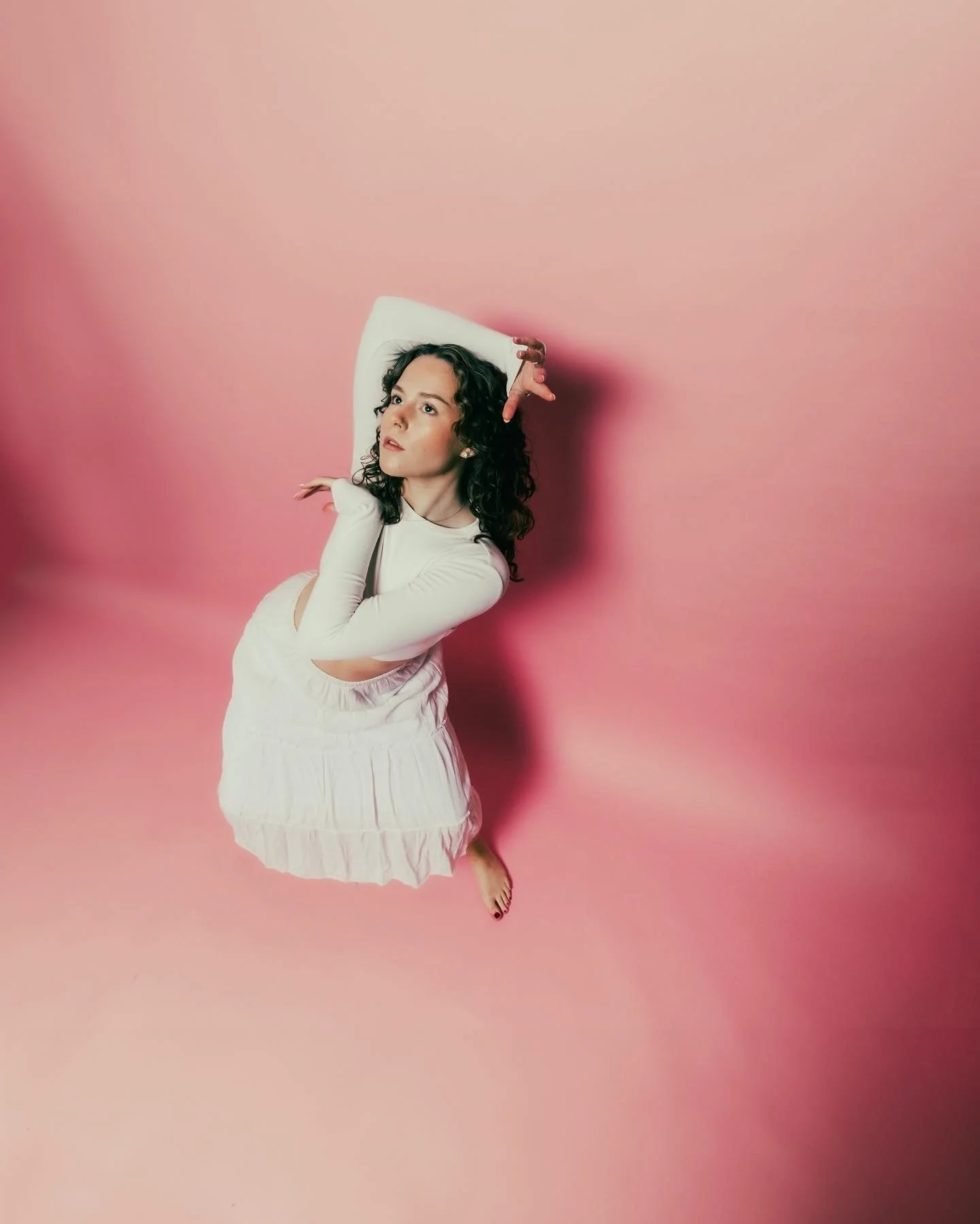A woman with curly dark hair, wearing a white long-sleeve top and a white skirt, laying on a pink background with her head resting on a pillow, looking thoughtful.