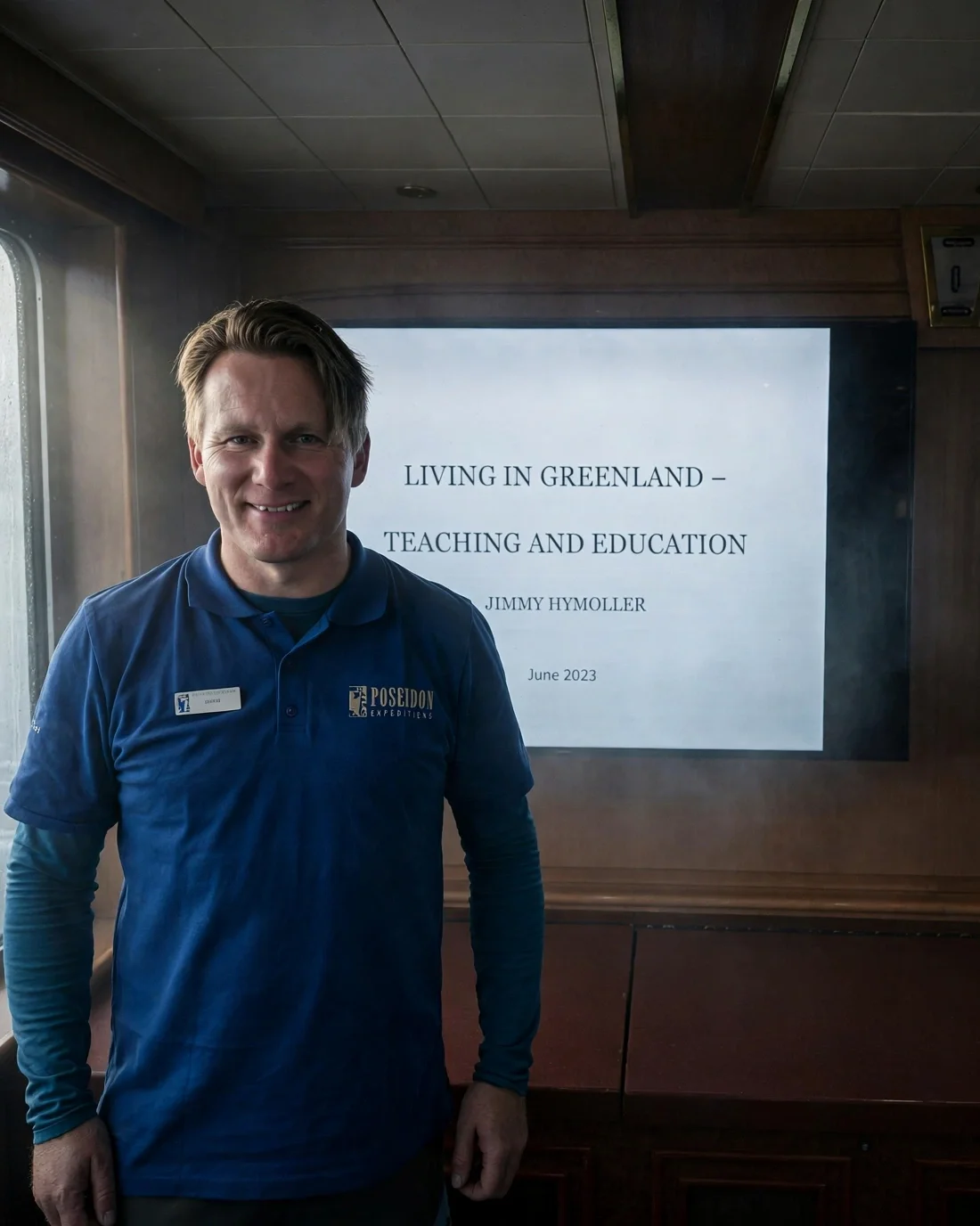 A man standing in front of a presentation slide titled 'Living in Greenland - Teaching and Education', presented by Jimmy Hymoller in June 2023, inside a wooden-paneled conference room, on a ship or boat, wearing a blue Poseidon Expeditions shirt and smiling.