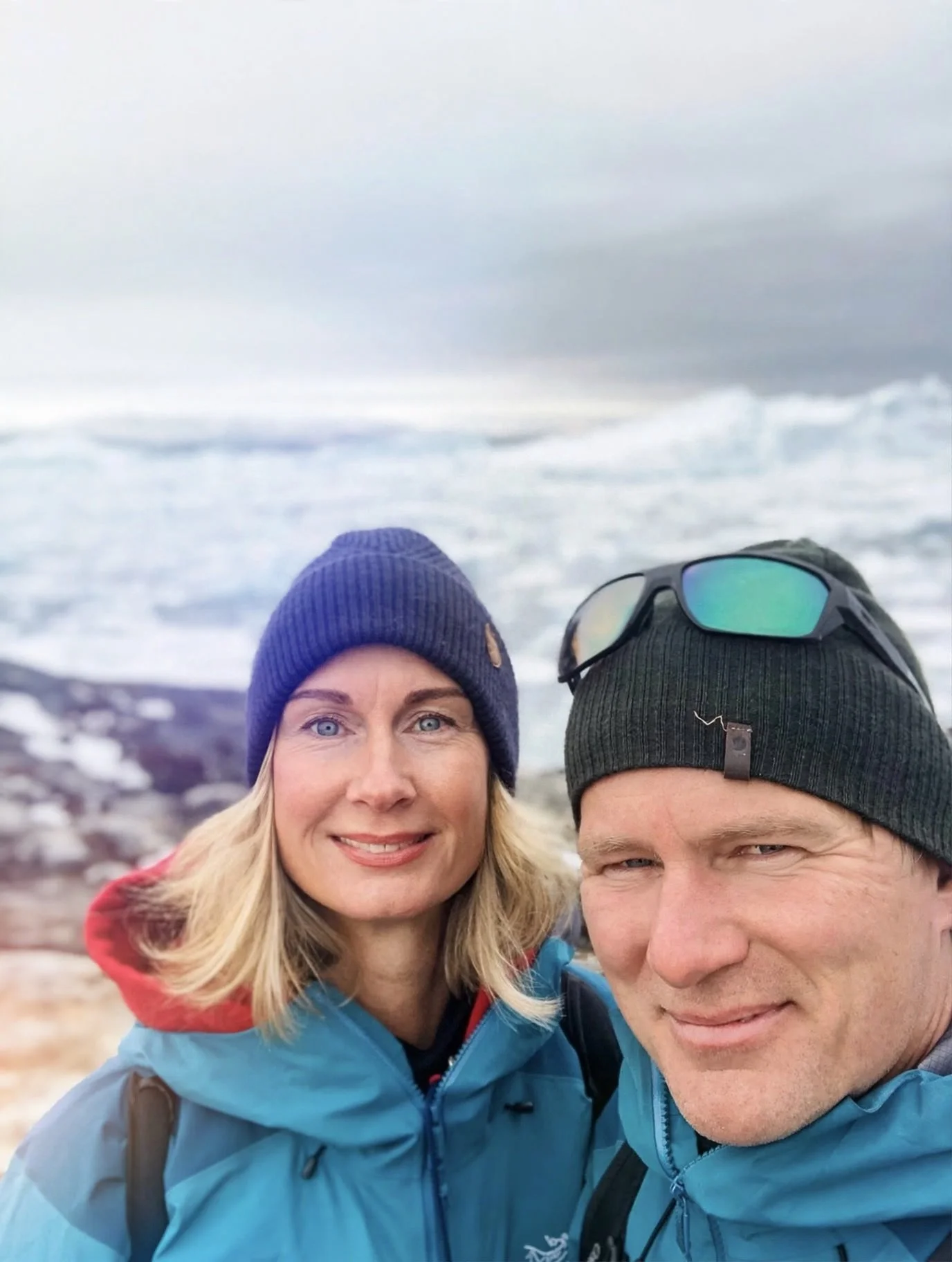 A woman with blonde hair and a blue beanie, and a man with brown hair, both smiling, in front of a shoreline with ocean waves, wearing outdoor jackets.