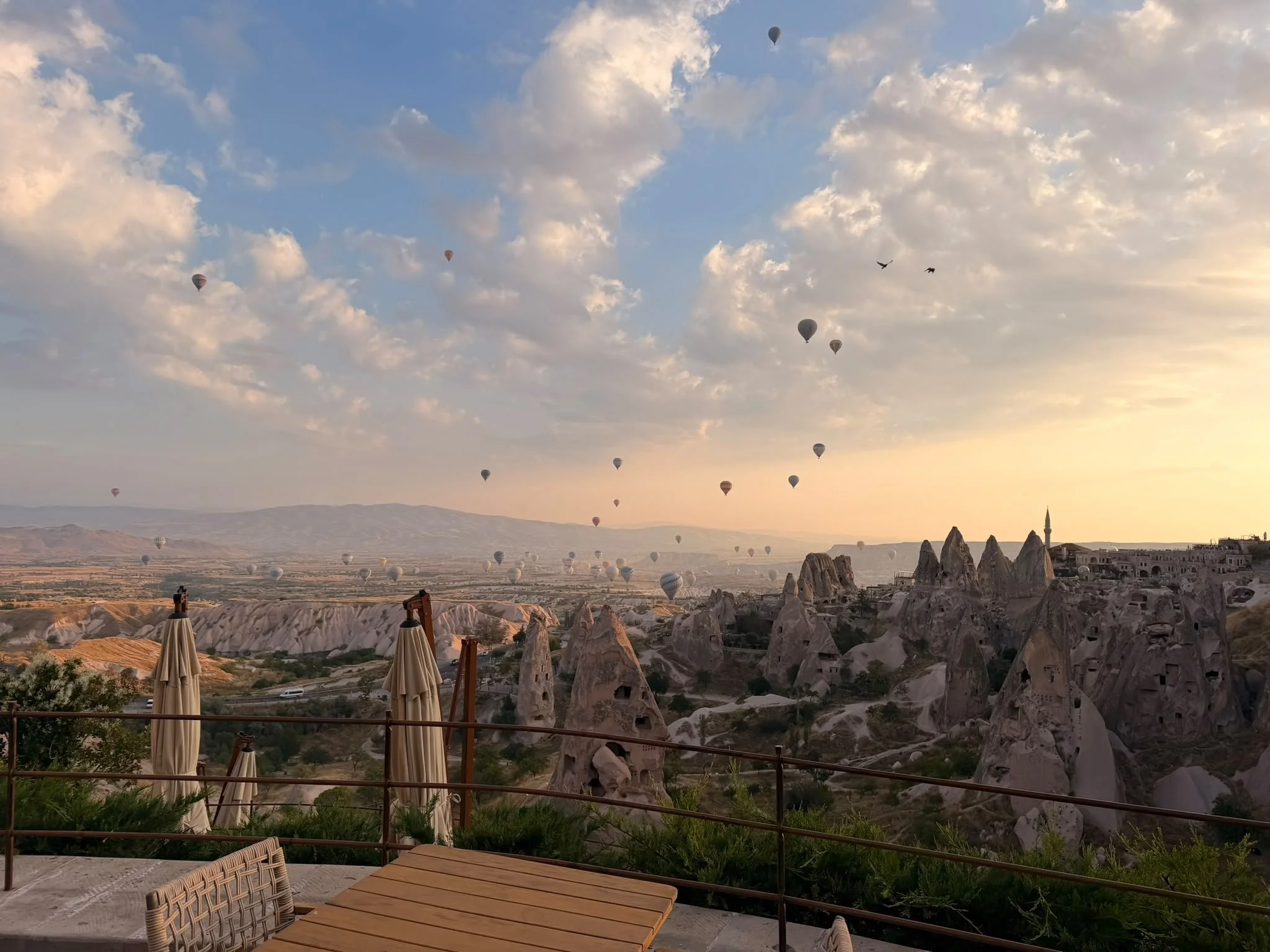 Blick auf den Himmel mit Heißluftballons über einer Felsenlandschaft bei Sonnenuntergang in Kappadokien, Türkei, mit Tafel und Stühlen im Vordergrund.