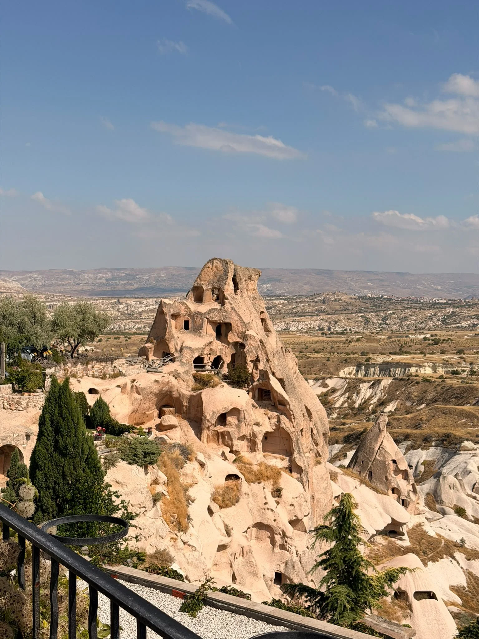 Felsenstadt in Kappadokien, Türkei, mit Höhlenwohnungen und Felsenformationen, grüne Bäume im Vordergrund, blauer Himmel mit wenigen Wolken, im Hintergrund Hügel und Ebene