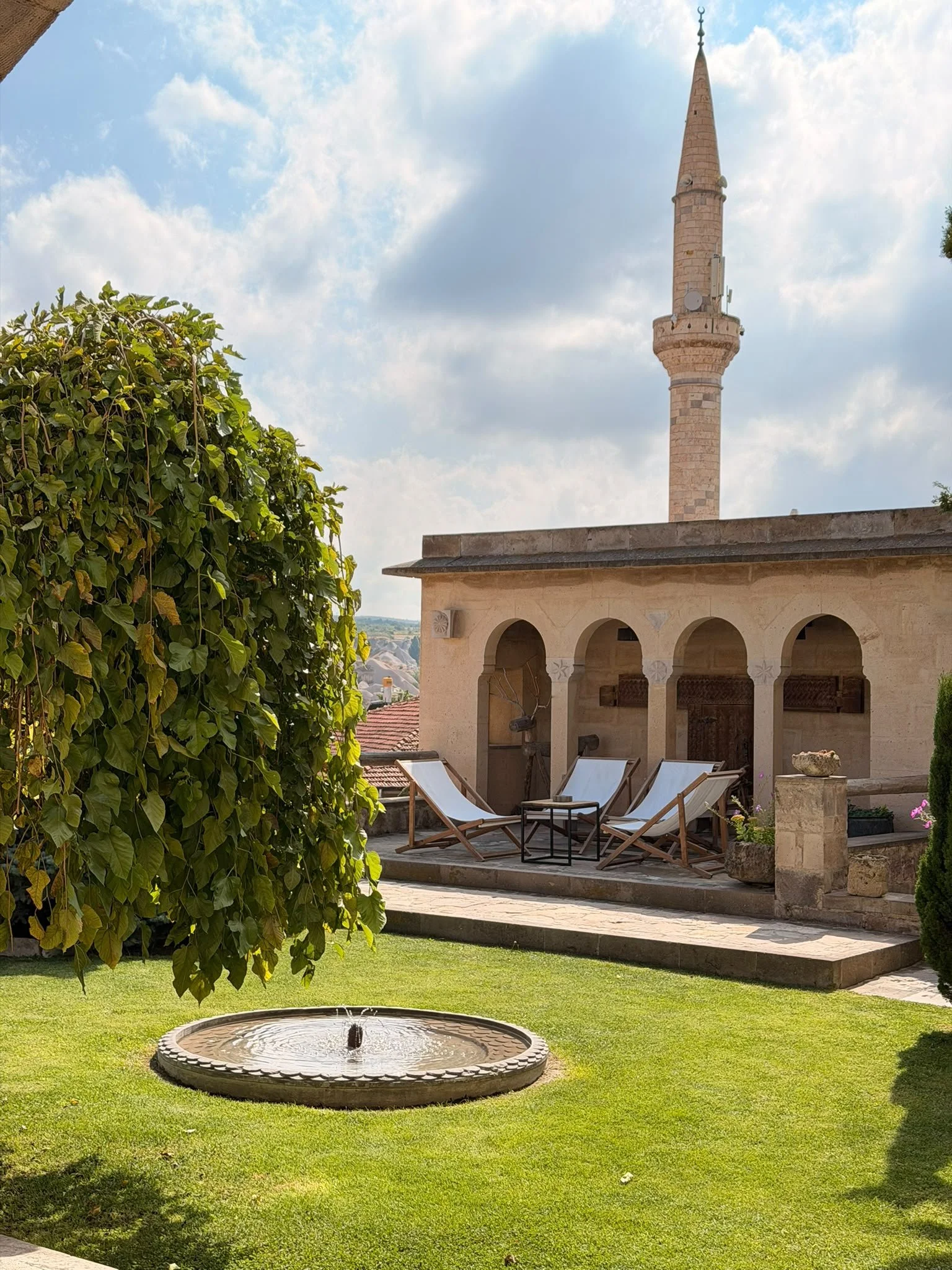Garten mit Brunnen im Vordergrund, Sitzgelegenheiten und eine Terrasse mit Bogen an einer historischen Gebäude mit Turm im Hintergrund, gegen bewölkten Himmel.