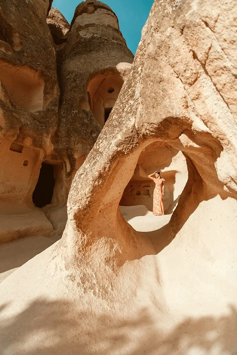 Frau in langem Kleid steht in einer Felsöffnung in einer Sandsteinlandschaft mit mehreren Höhlen.}
