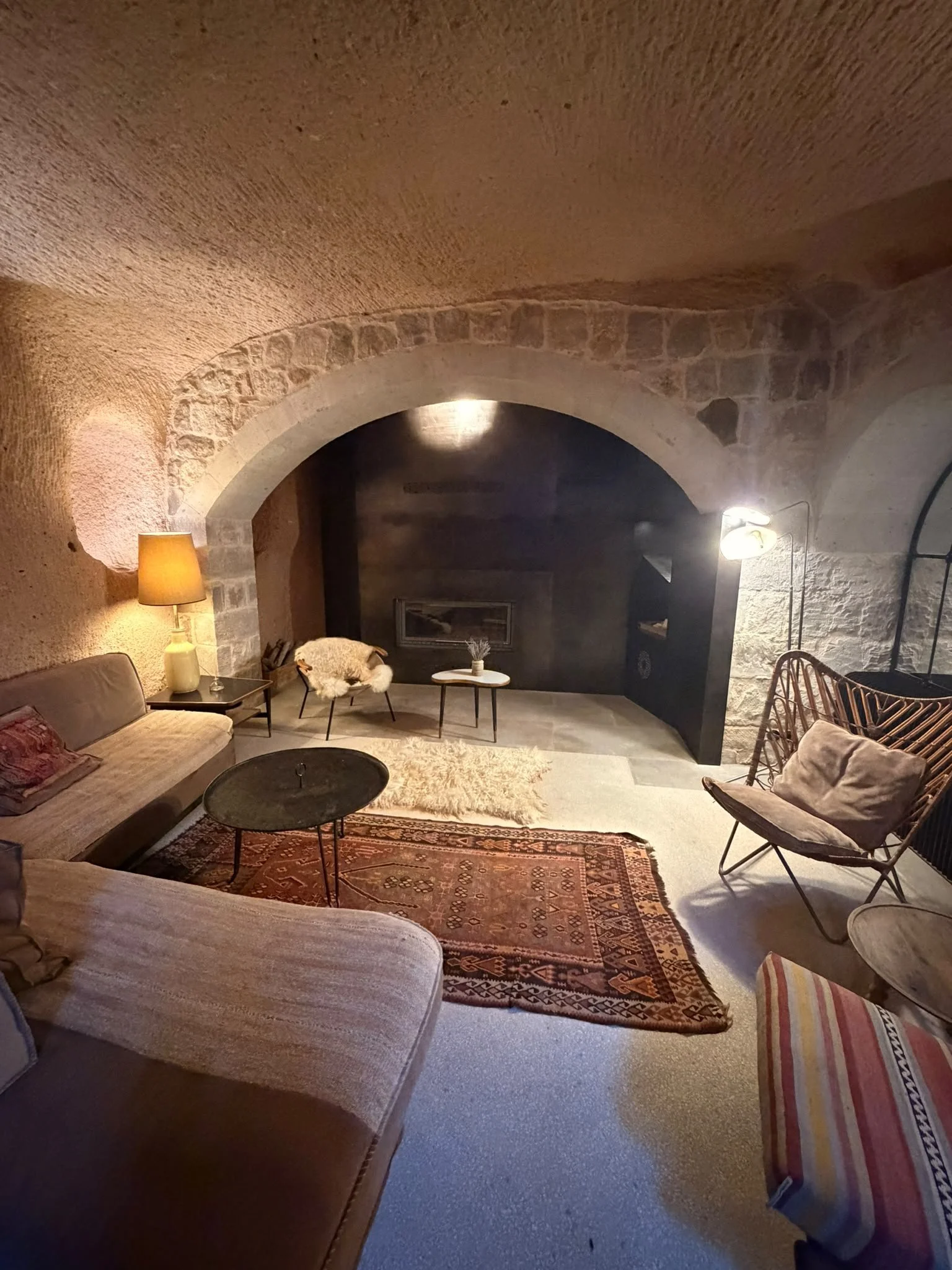 Gemütliches Wohnzimmer mit einem braunen Sofa, einem Teppich, einem Korbstuhl, einem Beistelltisch, einem Lampen und einer Steinbogenarchitektur an den Wänden.