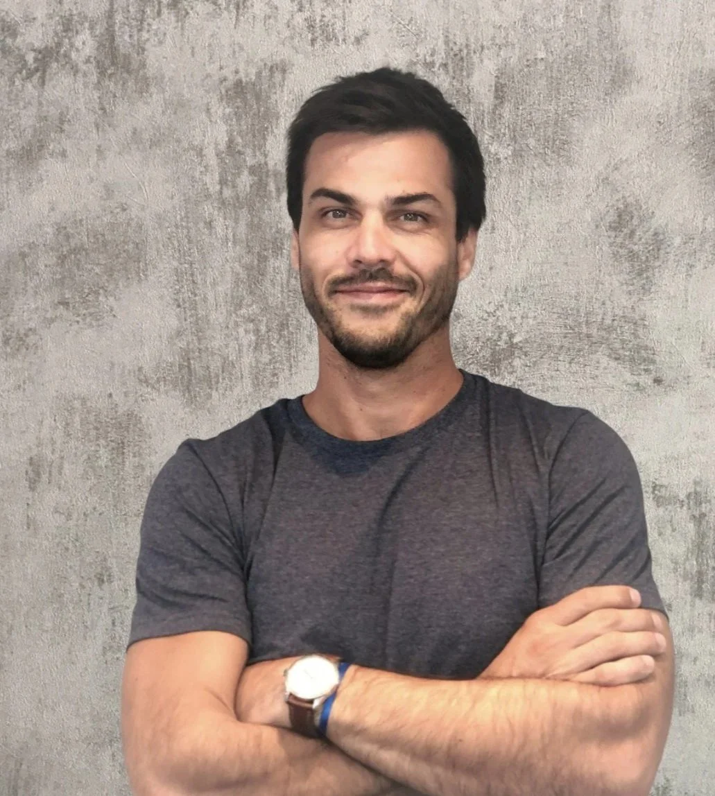 Un hombre con barba, cabello oscuro y camiseta gris, con brazos cruzados, sonriendo frente a una pared de concreto.
