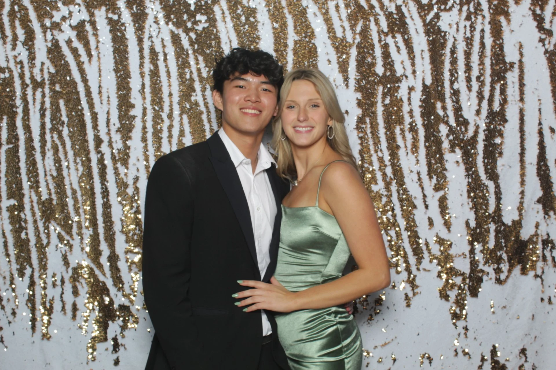 A young man and woman are dressed in formal attire, posing and smiling in front of a sparkling gold and white sequin backdrop at a celebration or prom.