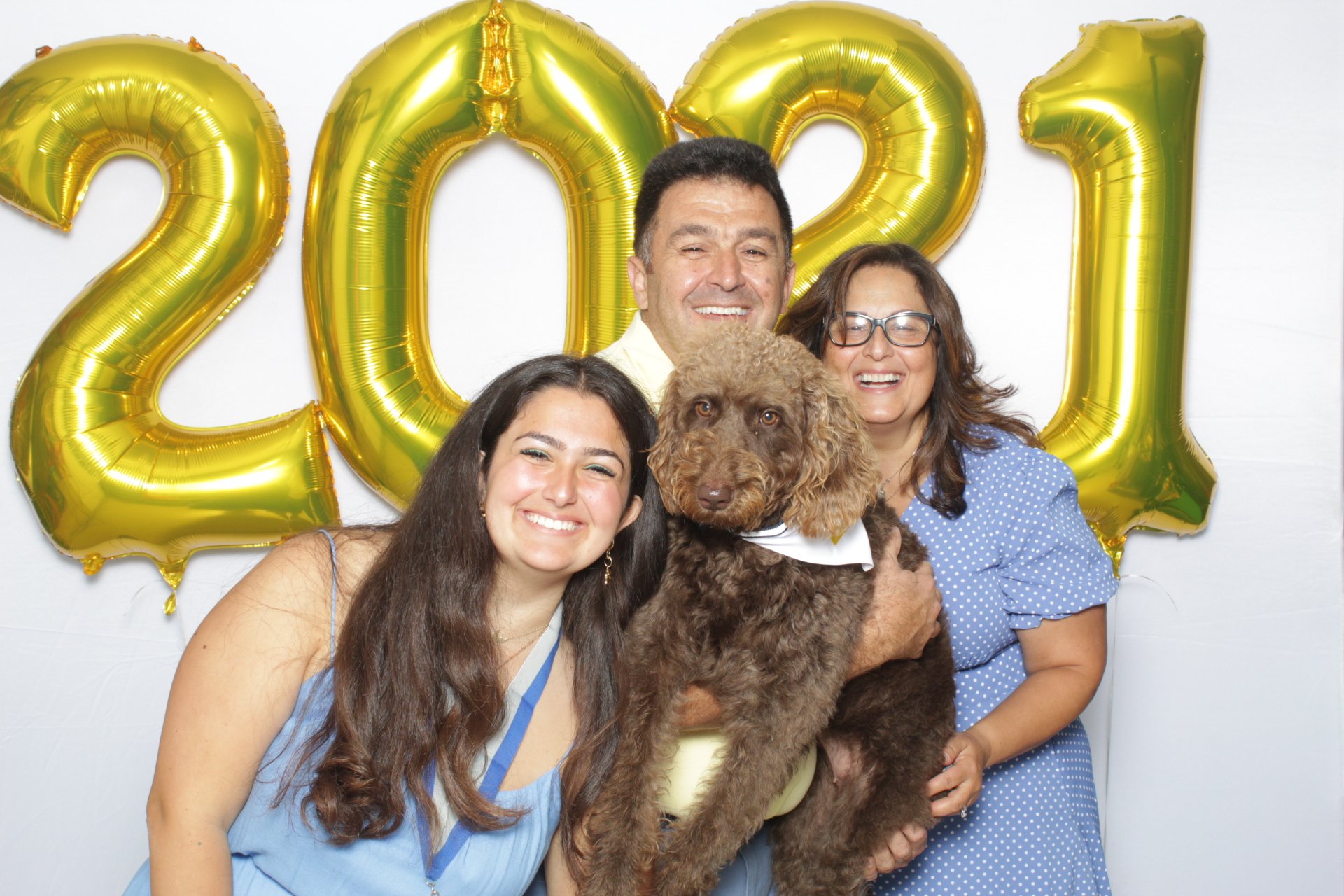A group of four people, two women and two men, standing together and smiling, holding a brown curly-haired dog. Behind them are large gold balloons that spell out '2021' on a white wall, indicating a celebration or New Year's event.