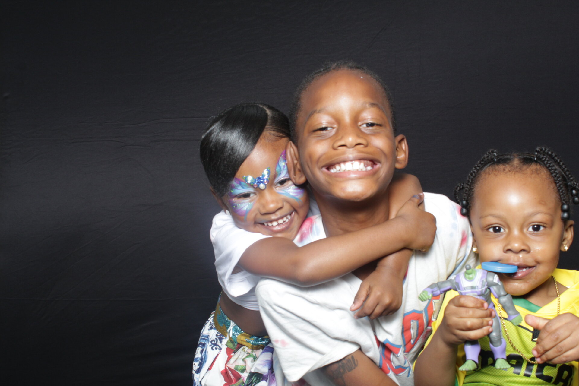 Smiling children embracing each other against a black background, with colorful face paint and toys.