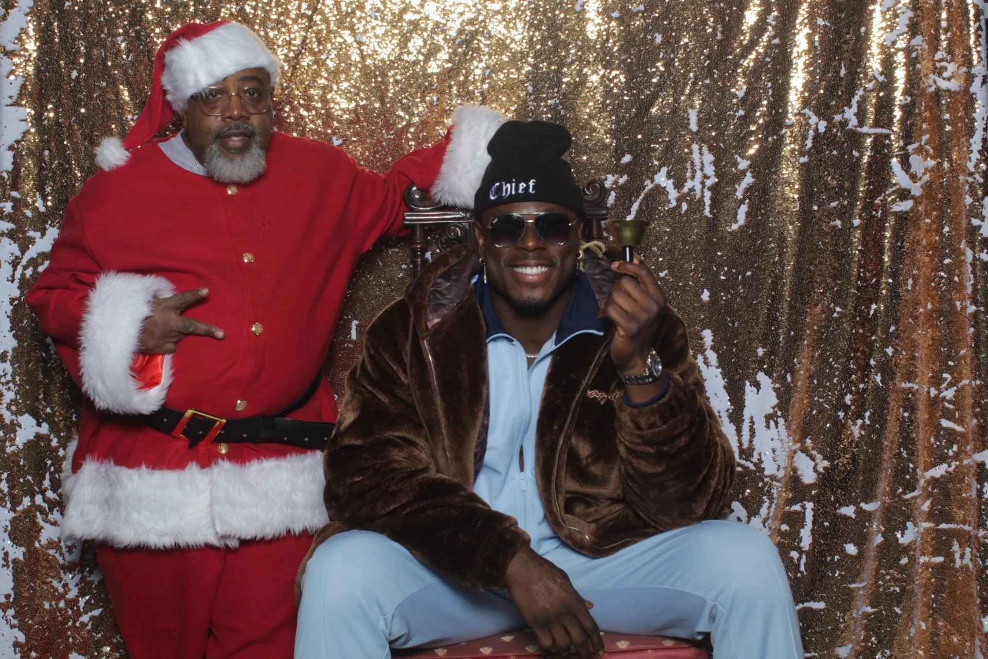 Two guests in Santa and festive attire posing against a gold sequin photo booth backdrop.