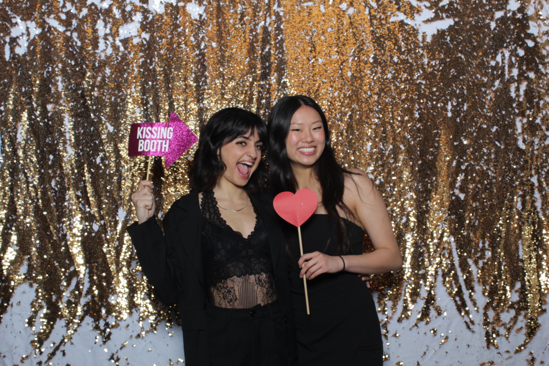 Two guests holding a heart and kissing booth props against a gold and white sequin backdrop.
