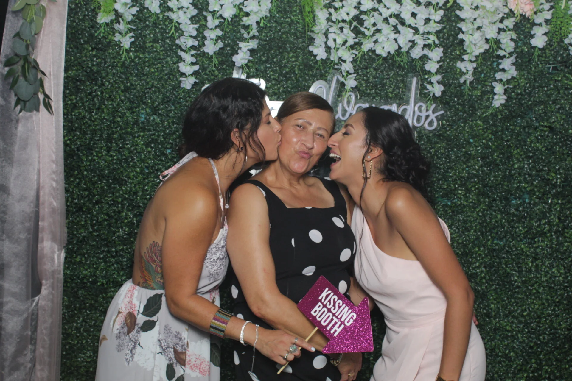 Two women kissing an older woman on the cheeks at a wedding, with a green leafy wall background and a sign that reads 'Kissing Booth'.