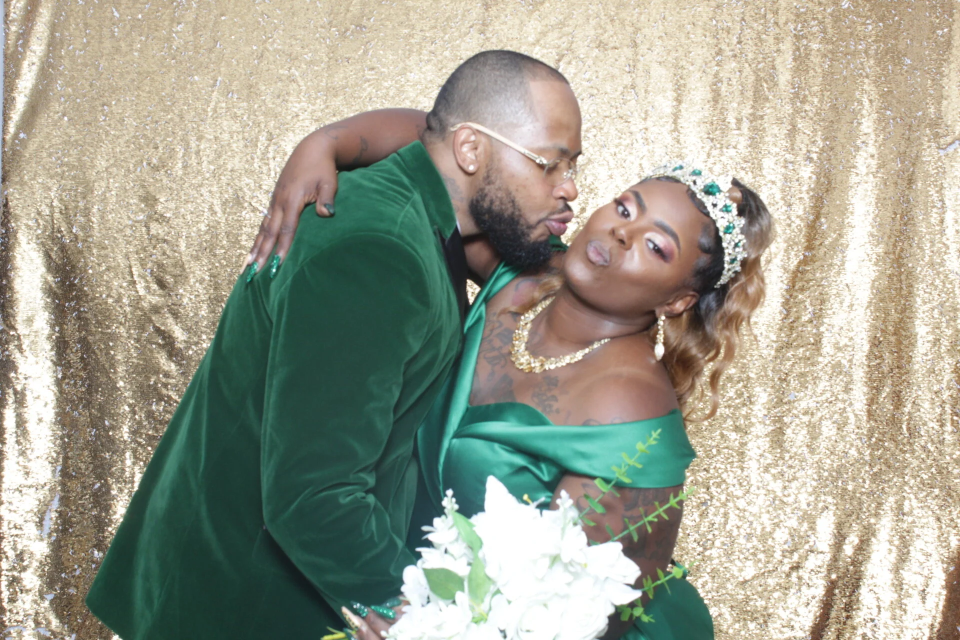 A couple dressed in matching green attire, posing close together in front of a gold sequin backdrop, with the woman holding a bouquet of white flowers and wearing jewelry and a tiara, while the man wears glasses and has a beard.