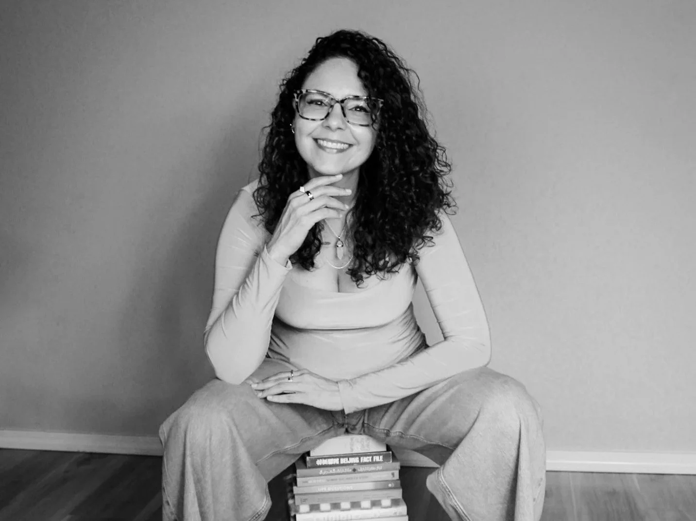 Black and white photograph of a woman with curly hair wearing glasses, a fitted long-sleeve top, and loose jeans, sitting on a stack of books against a plain wall, smiling at the camera.