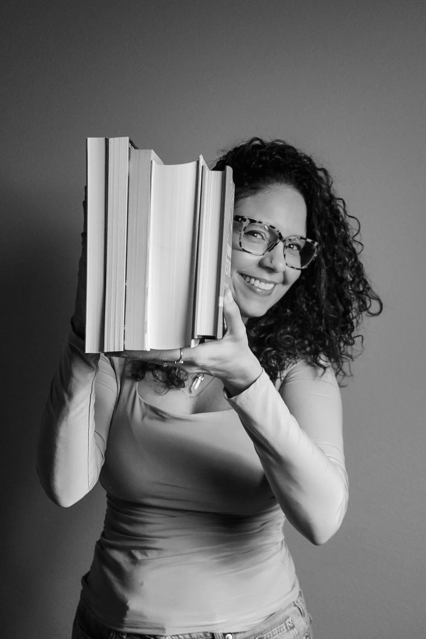 A woman with curly hair and glasses is smiling while holding a stack of books with one eye peeking out from behind the books. She is wearing a long-sleeved shirt and pants, and standing against a plain background.