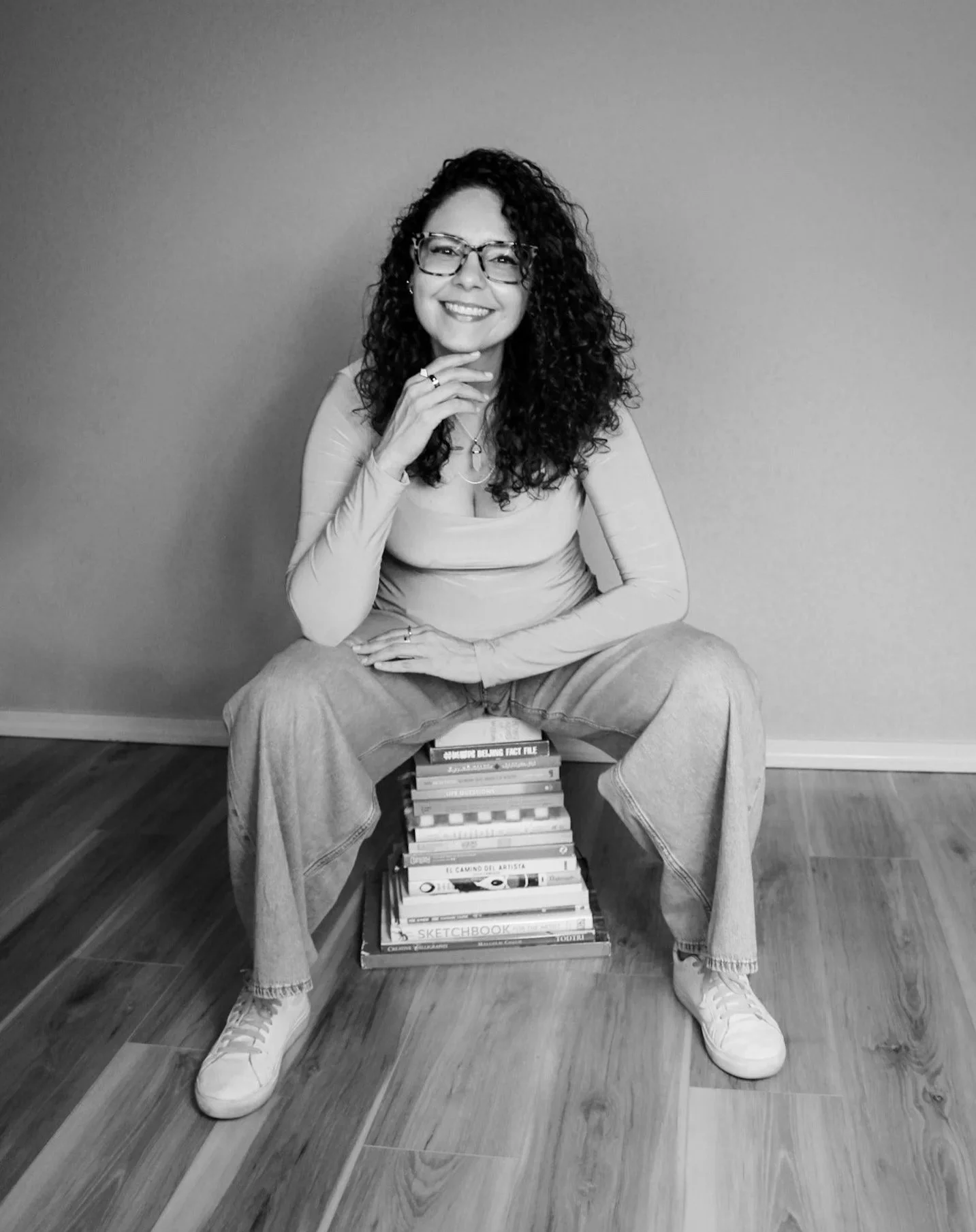 Black and white photo of a woman with curly hair and glasses sitting on a stool, smiling, with her hand on her chin. She is wearing a long-sleeve top, loose jeans, and sneakers. There is a stack of books underneath the stool, and the room has a hardwood floor and plain wall background.