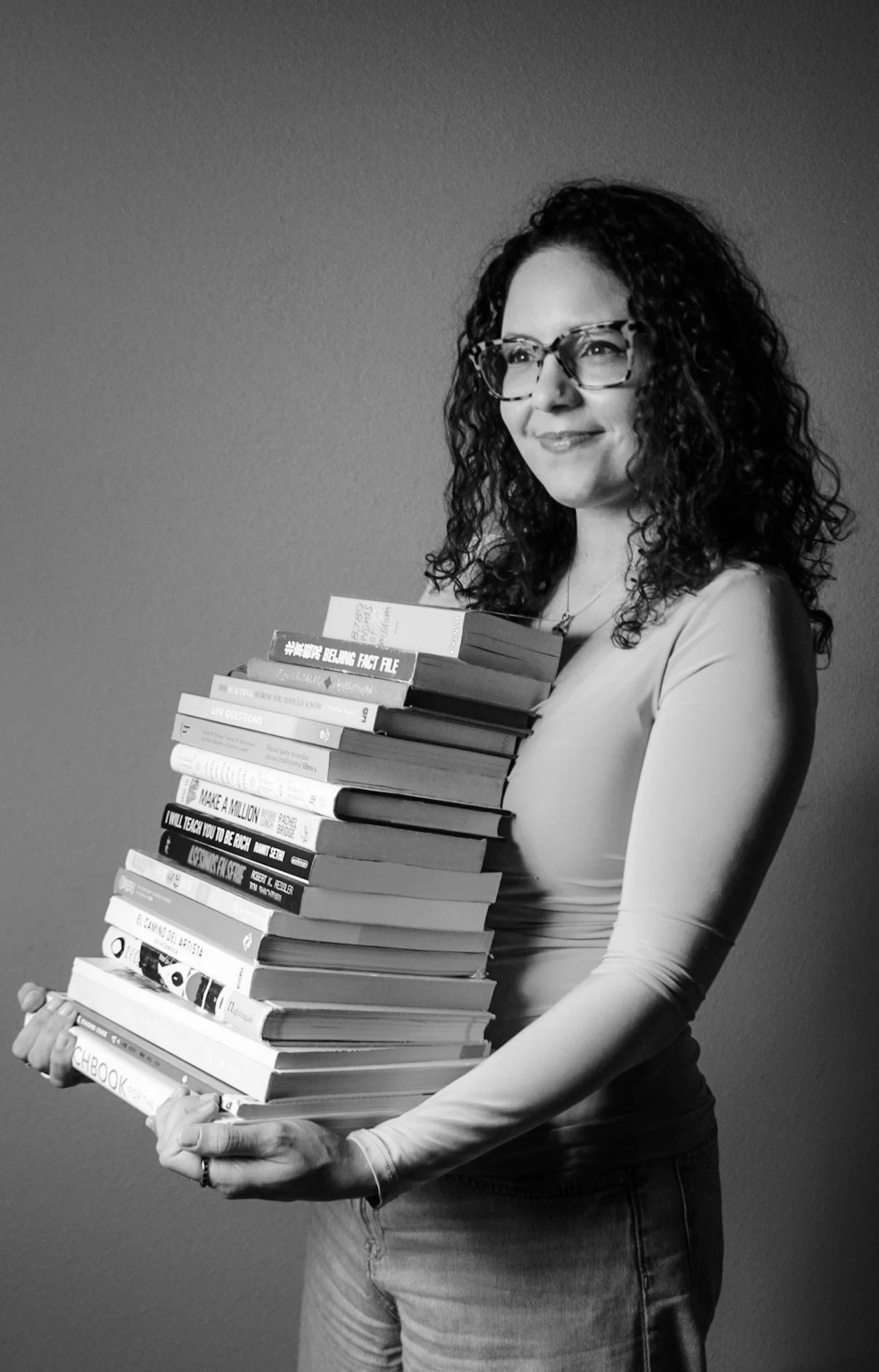 A woman with curly hair and glasses holding a tall stack of books, smiling, in a black-and-white photo.