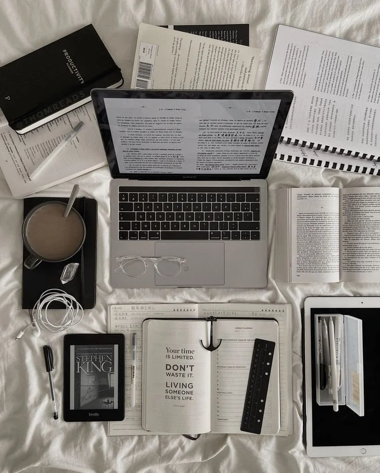 A flat lay of a workspace with a laptop, tablet, e-reader, numerous open books and papers, a cup of coffee, a pair of glasses, a ruler, pens, and headphones on a white surface.