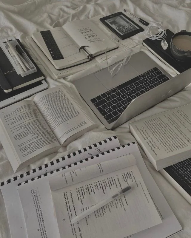 An organized study workspace with open books, a laptop, a planner, a pen, a smartphone, earphones, a cup of coffee, and stationary on a bed with white sheets.