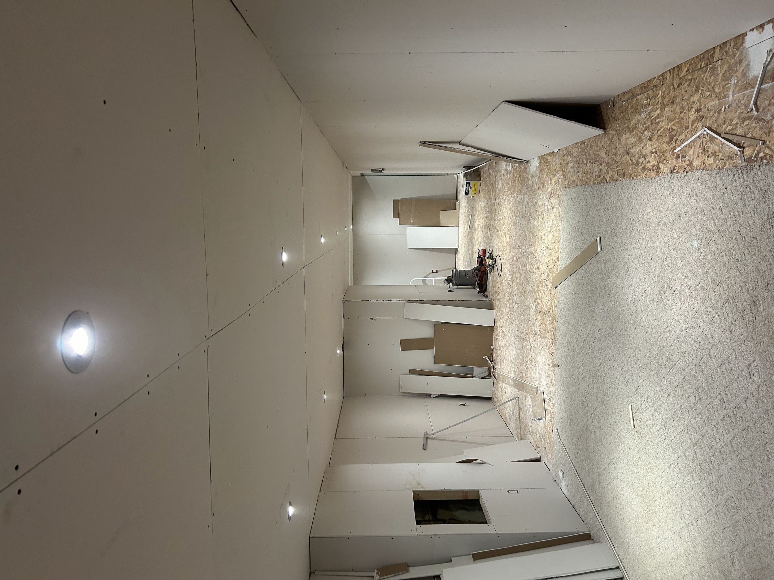 Interior of a building under renovation with partially finished walls, recessed ceiling lights, and construction materials and tools scattered on the floor.