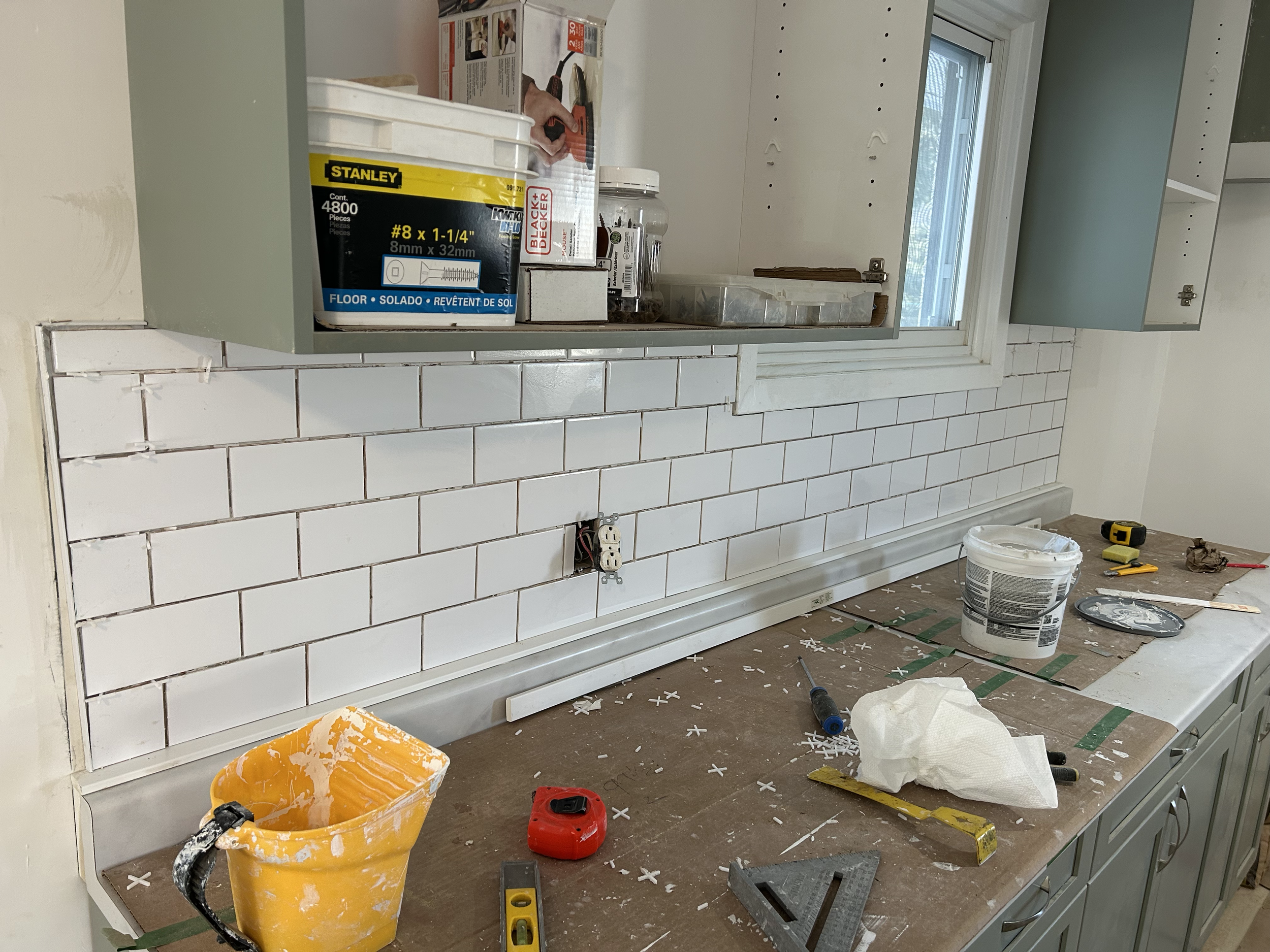 A partially renovated kitchen countertop area with tools and supplies scattered around. There are white subway tiles on the wall, a box of screws labeled 'Stanley', a hand holding a tool, a glass jar, a yellow bucket, a tape measure, and other constr