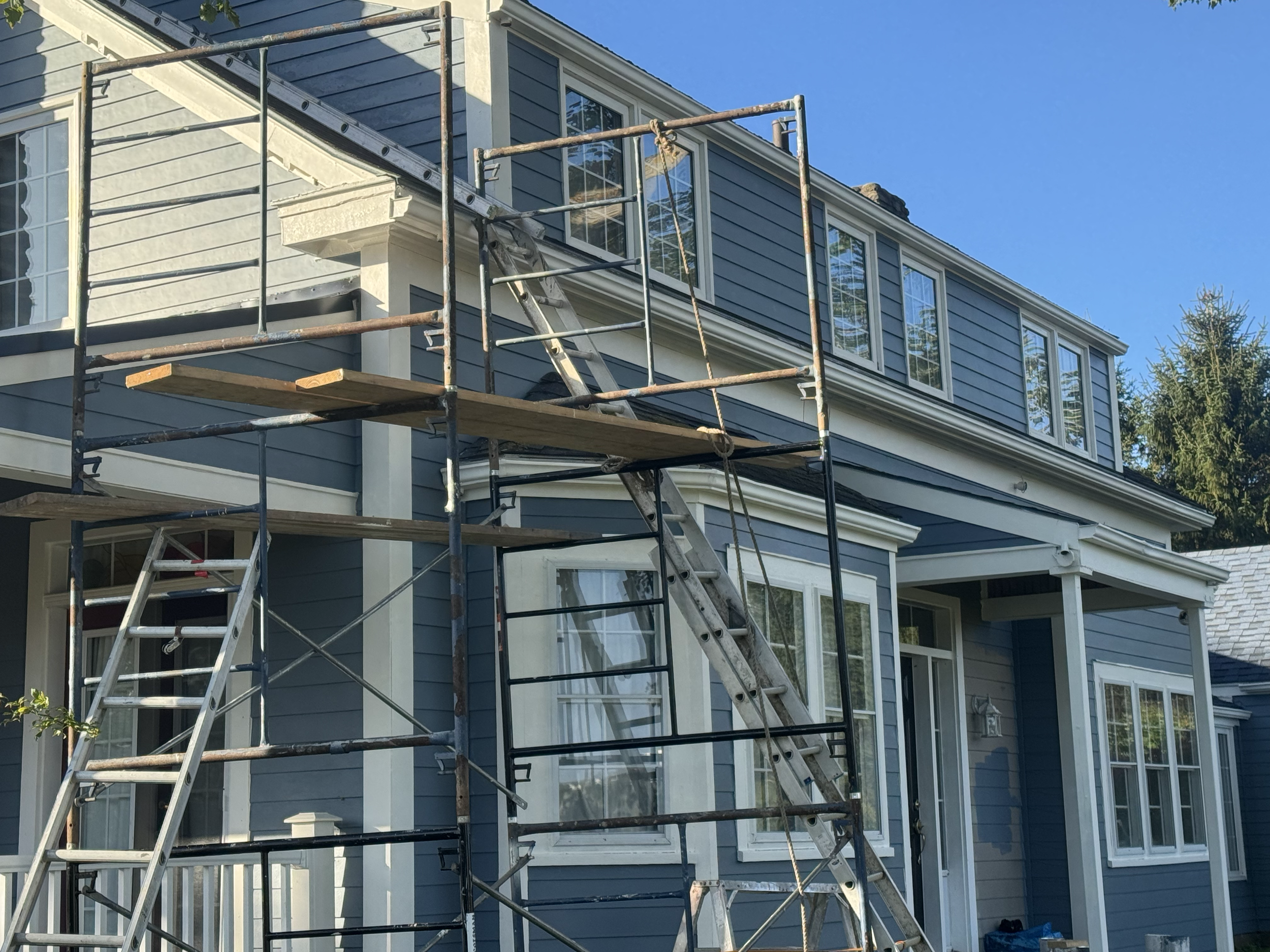 The image shows a house with blue siding and multiple windows. There is a metal scaffold with wooden planks set up on the side of the house, indicating ongoing exterior construction or renovation work. The sky in the background is clear and blue, and