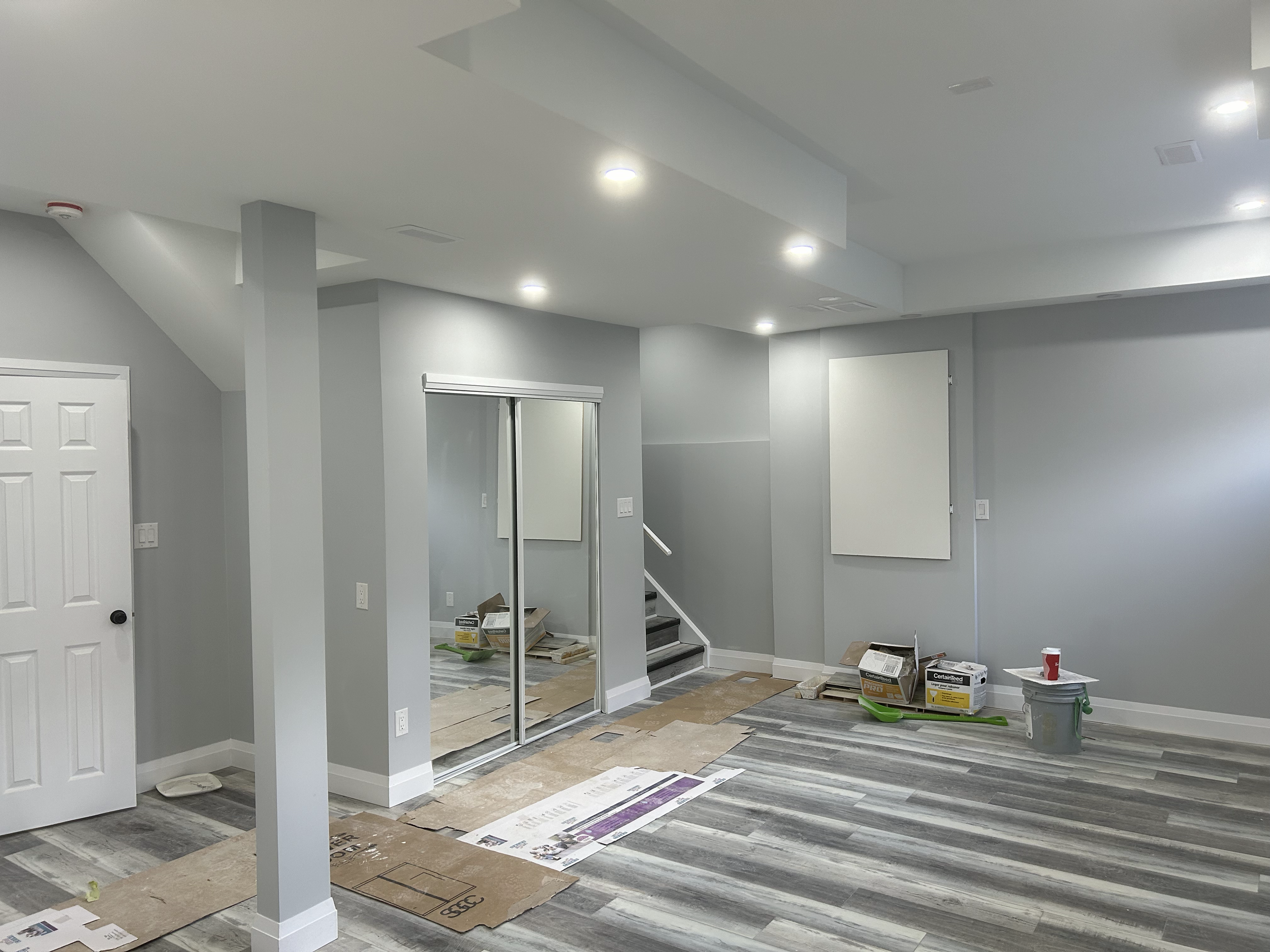 A room under renovation with floors covered in protective paper and tools scattered around. The walls are painted light gray and there are recessed ceiling lights. A mirror and boxes of construction materials are visible.