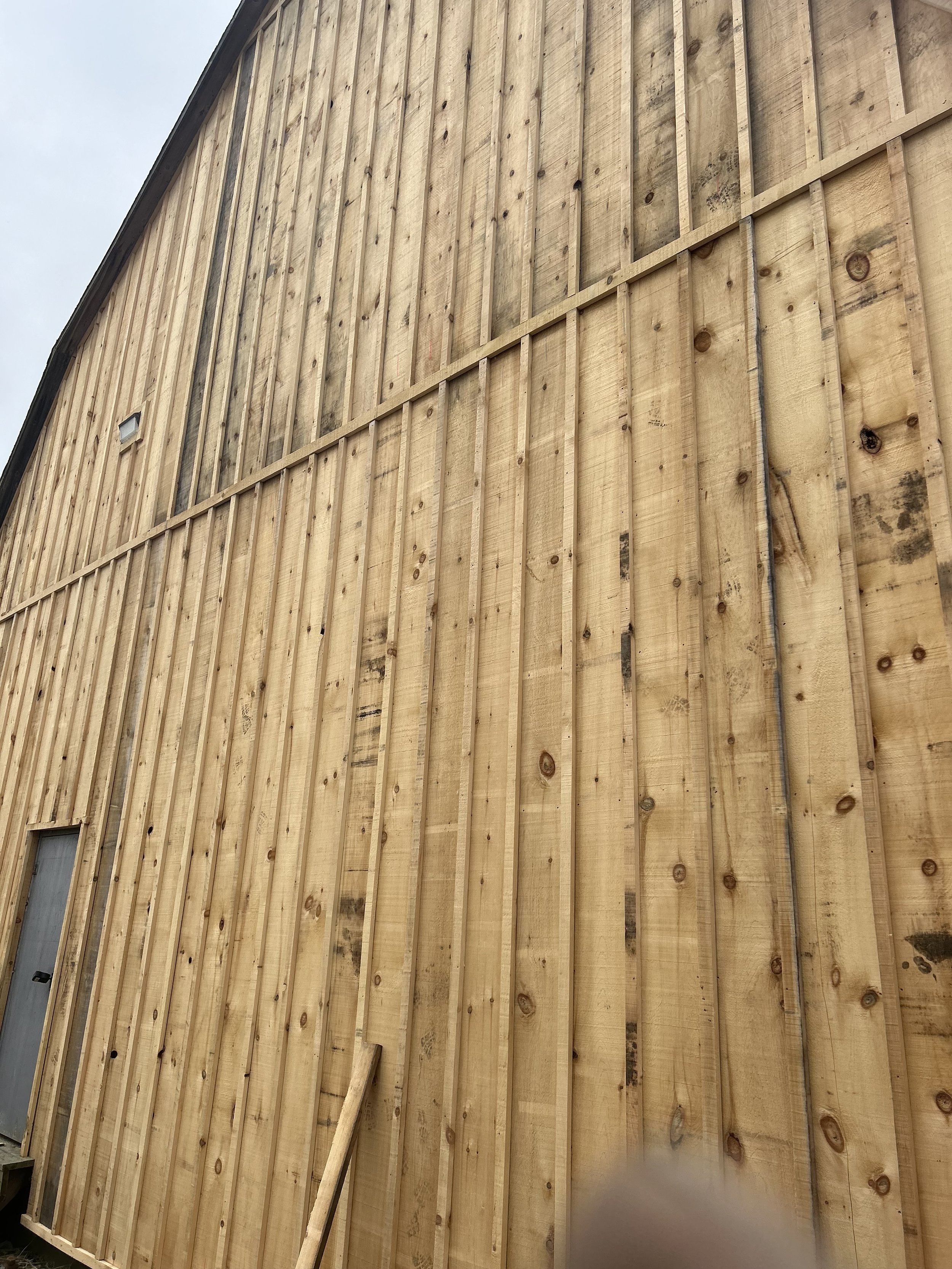 A large wooden wall under construction with vertical wooden planks, some leaning against the wall, and a small window near the bottom left corner.