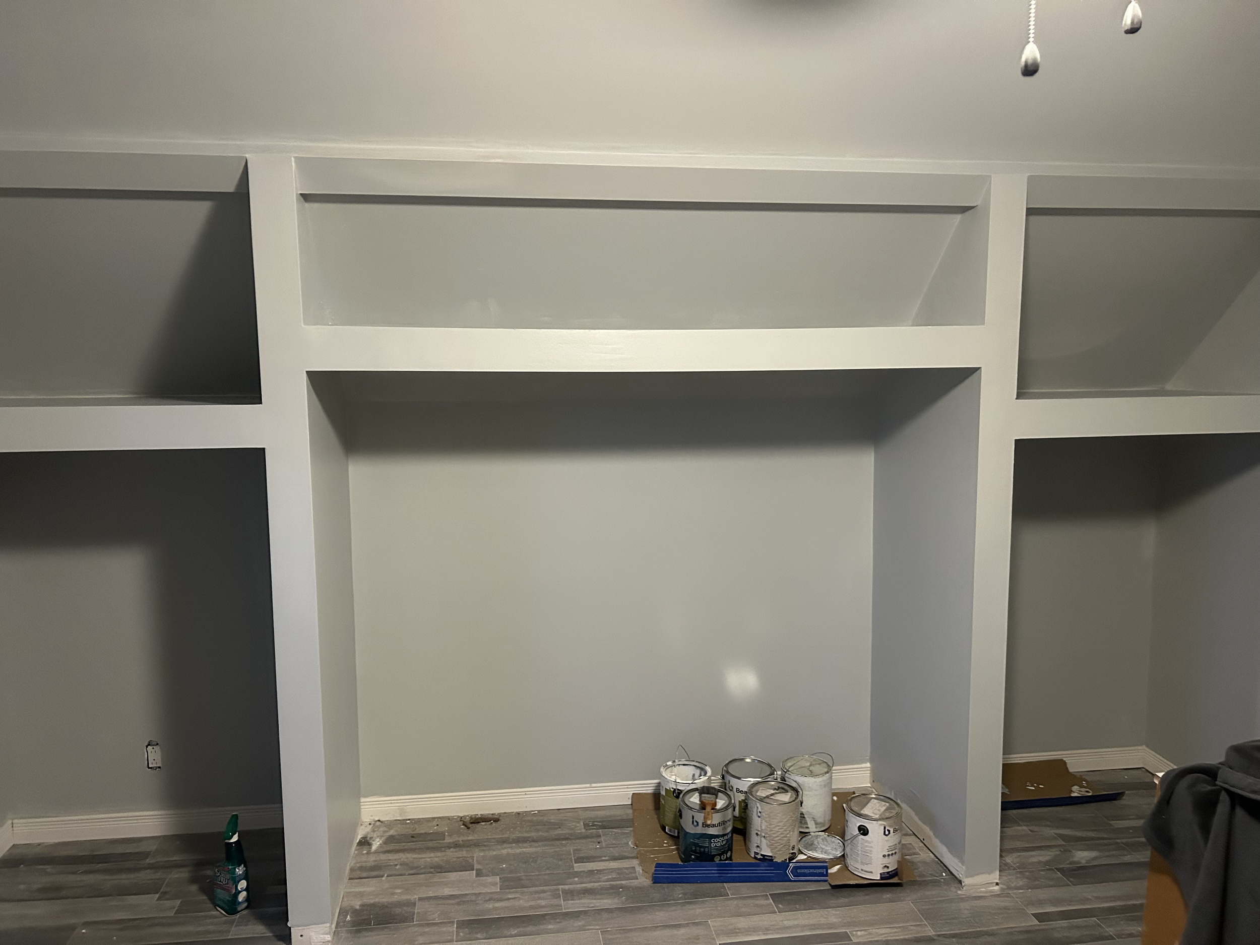 A partially assembled white shelf unit with three shelves, several cans of paint underneath, and painting supplies nearby on the floor. The room has gray walls and dark wood flooring.